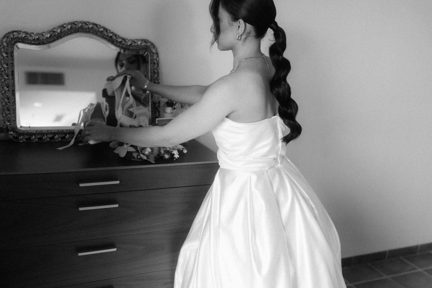 Black and white photo of a young girl in a white dress standing at a dresser with a mirror.