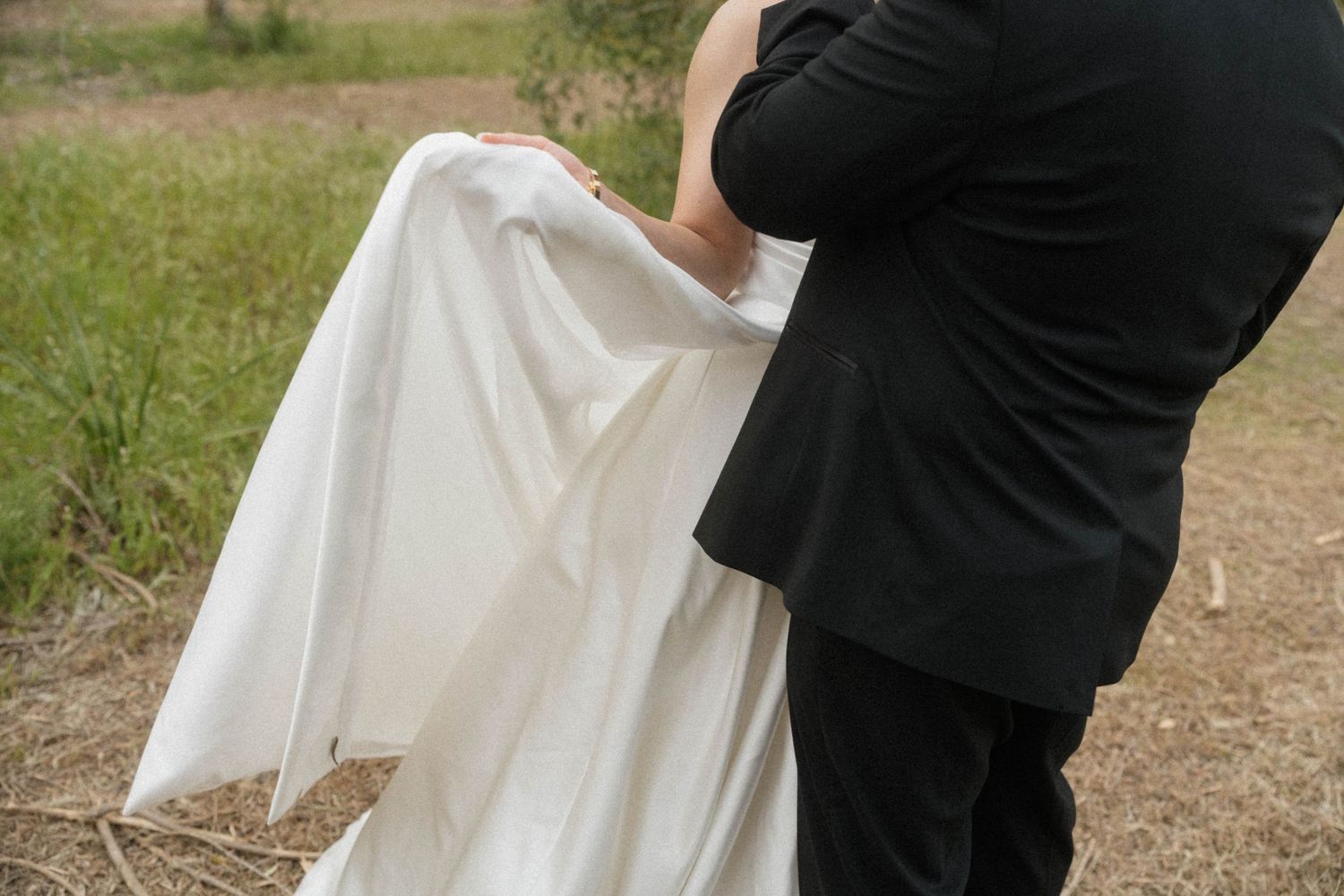 Groom in black suit tenderly holding bride's white wedding dress train outdoors.