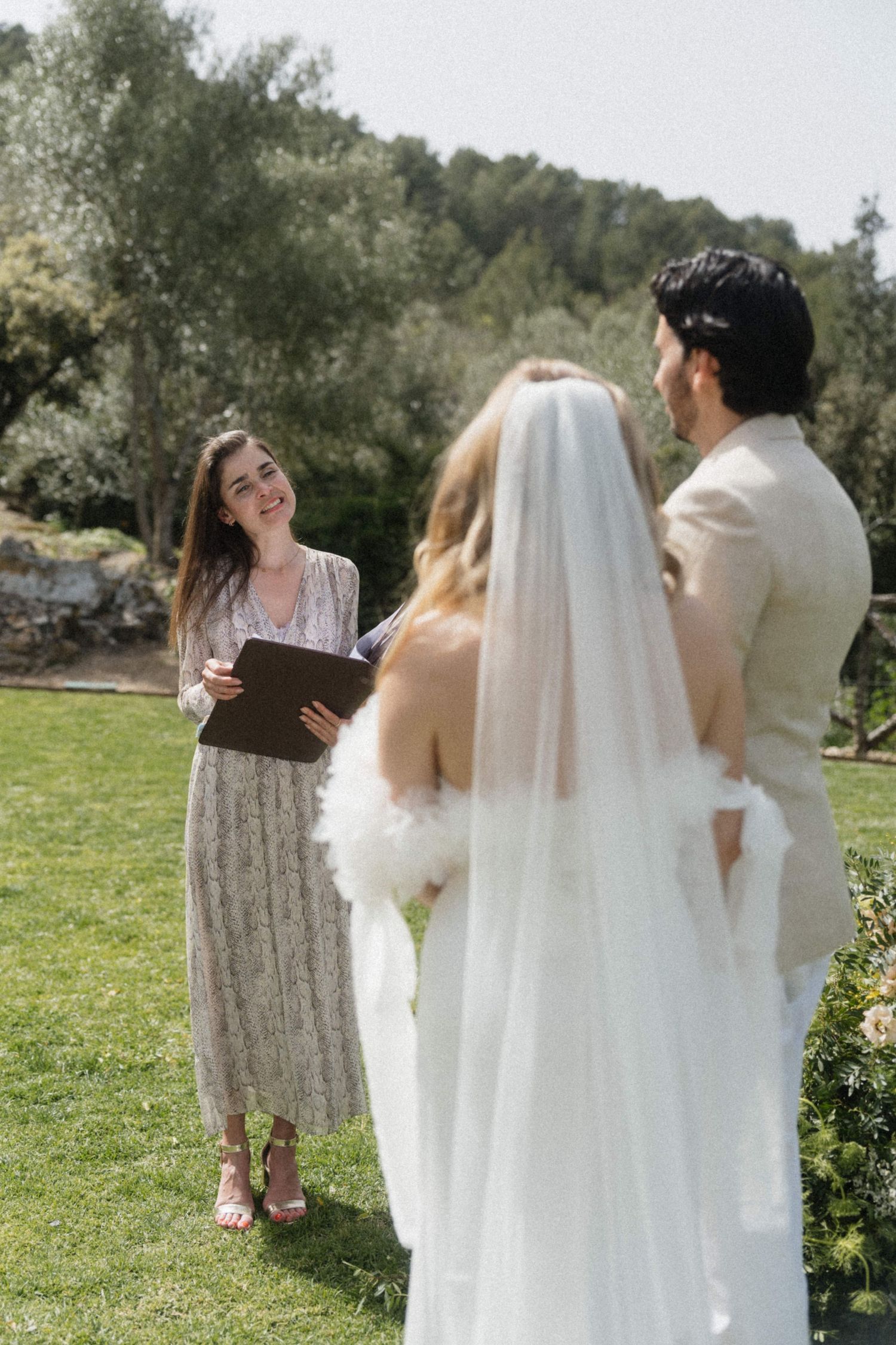 Smiling officiant reads vows to bride and groom during outdoor wedding ceremony on green lawn.