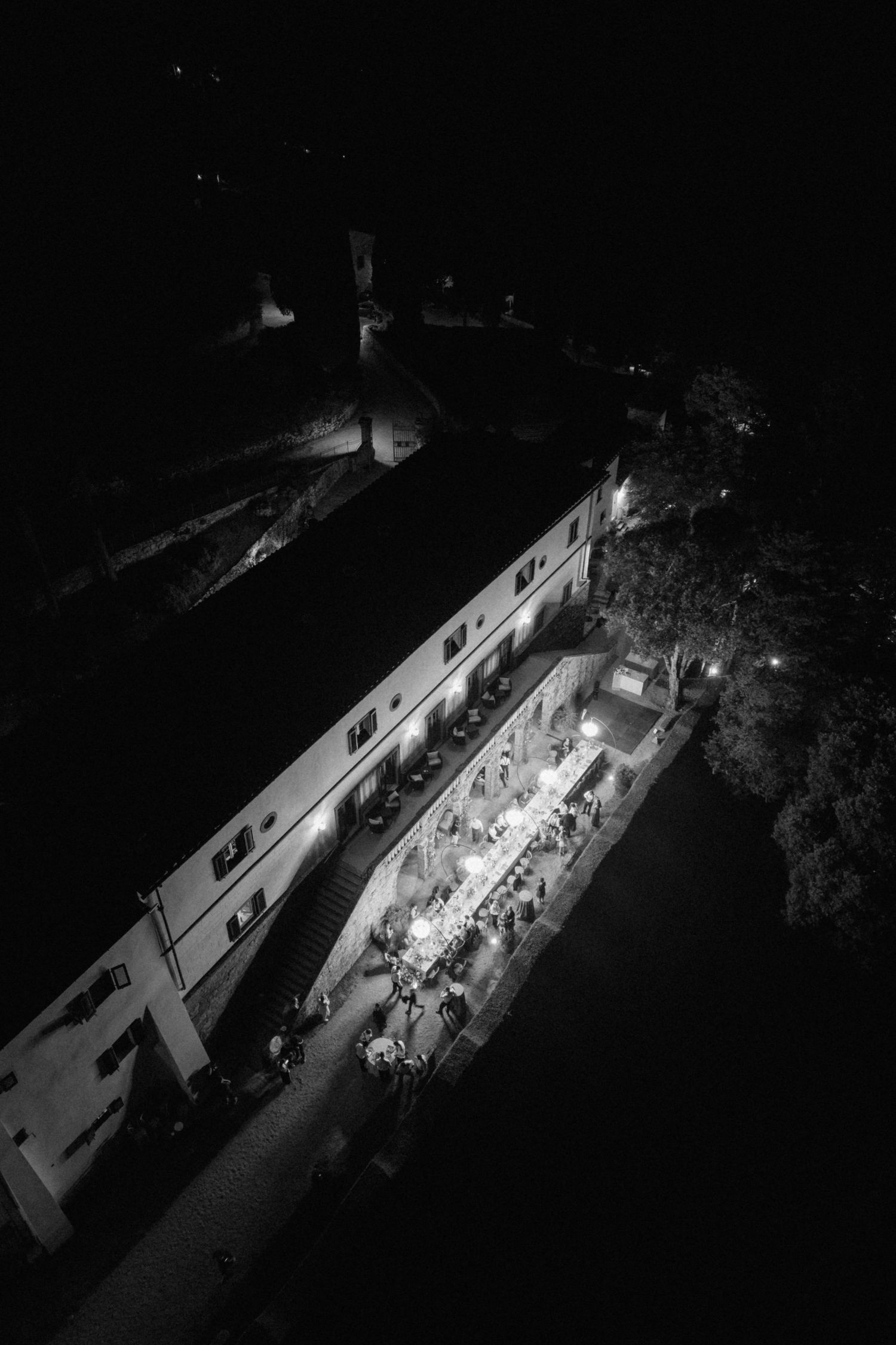 Aerial black and white night view of illuminated wedding reception venue with guests dining outside.