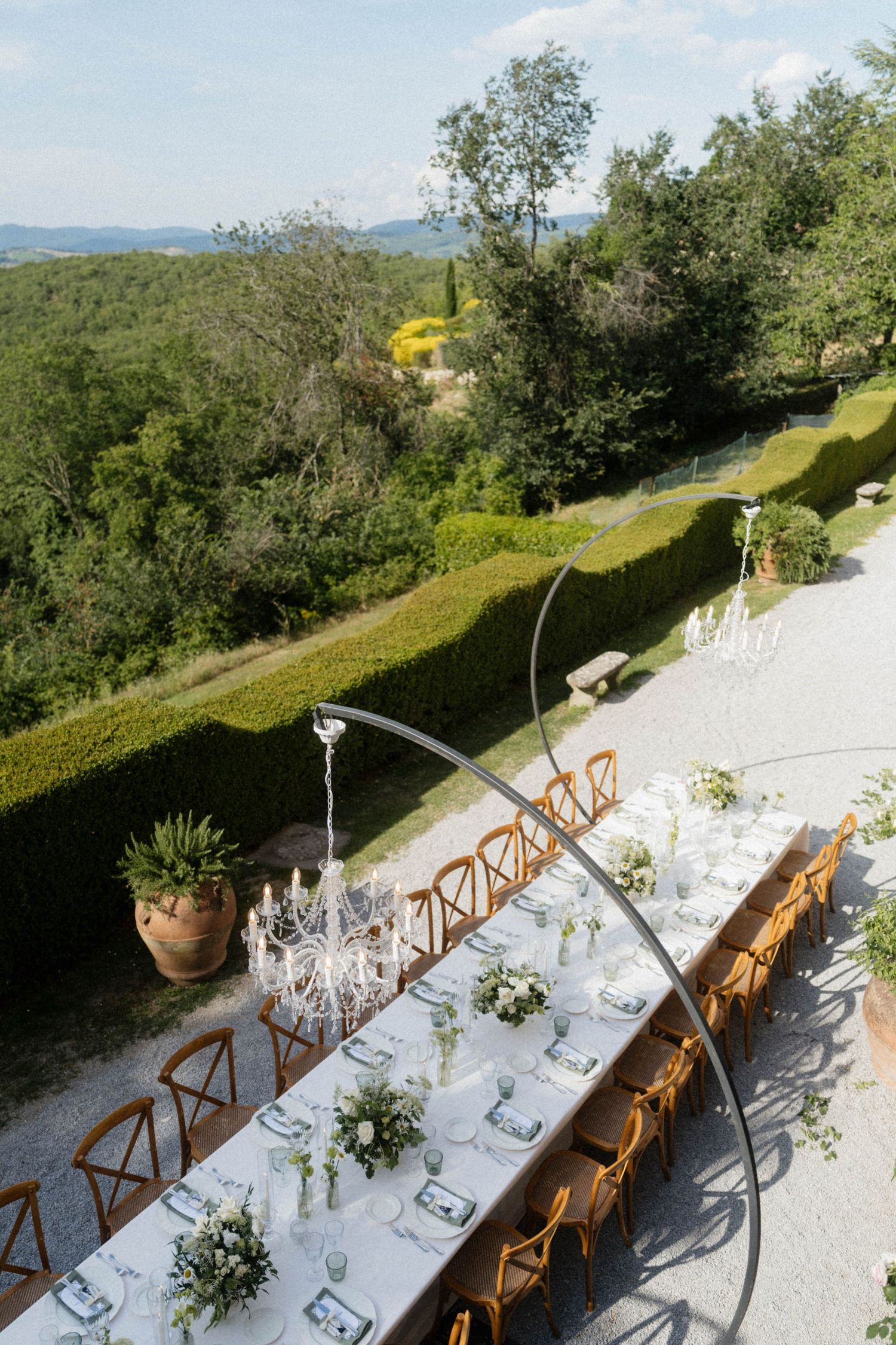 Breathtaking outdoor wedding reception table set against a lush Tuscan hillside landscape with crystal chandeliers.