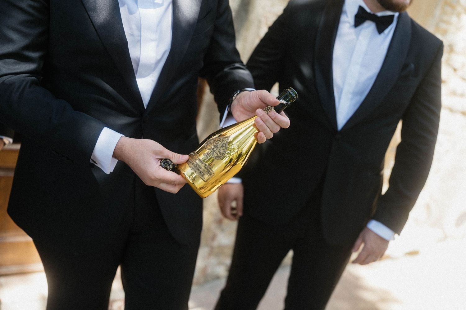 Two grooms in black tuxedos holding a gold champagne bottle at an outdoor wedding celebration.