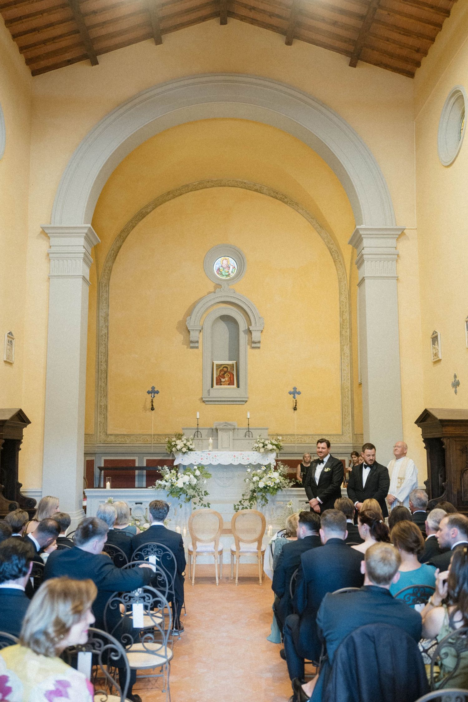 Wedding ceremony in Italian chapel with groom waiting at altar as guests fill wooden pews.