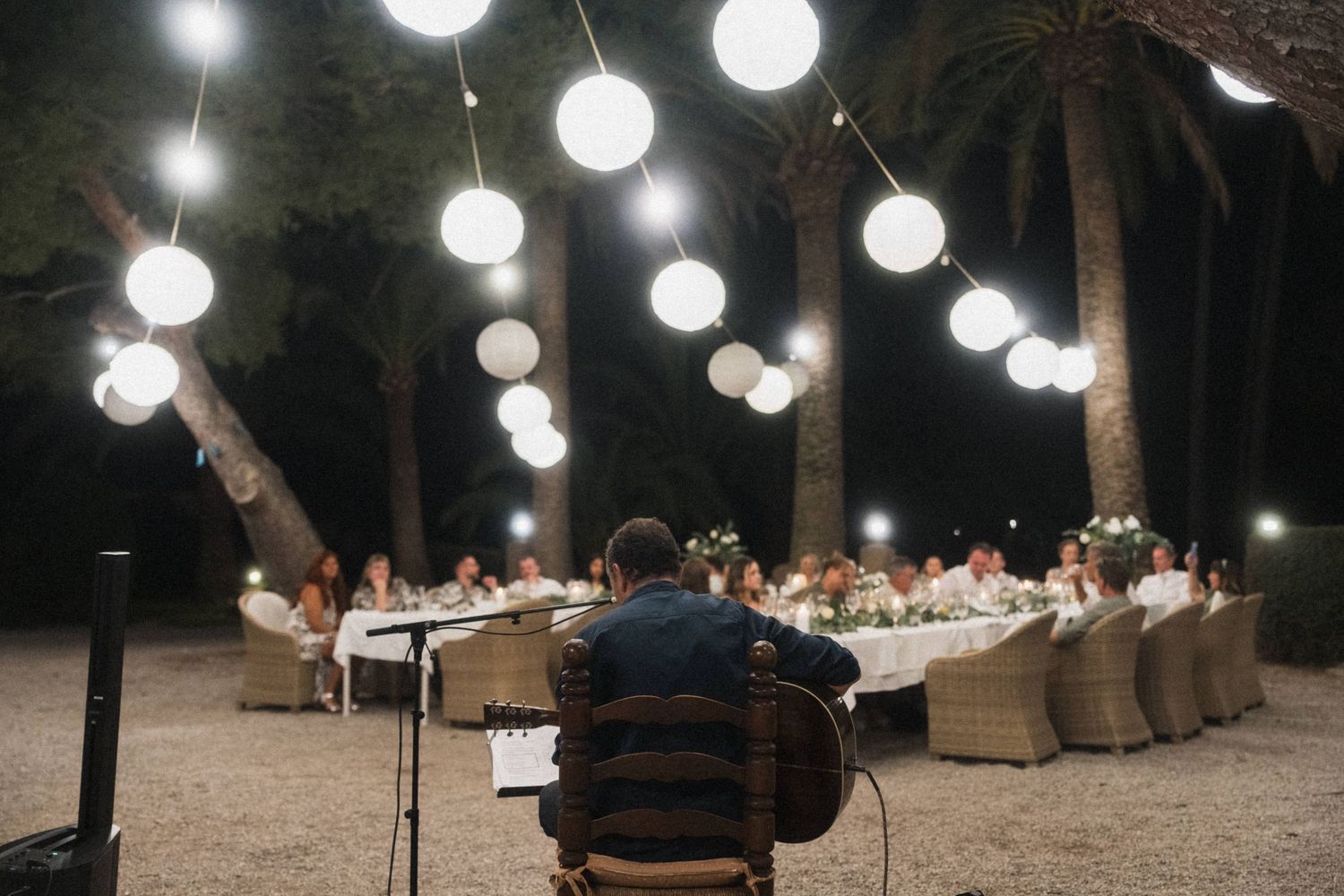 Outdoor evening wedding reception under glowing globe string lights with guests seated at long table among palm trees.