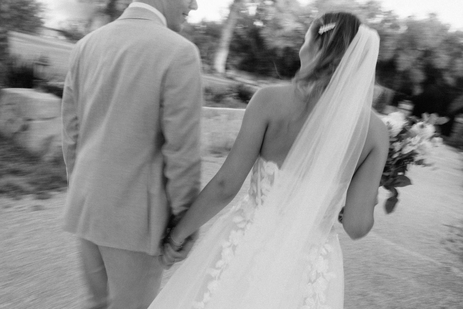 Black and white photo of bride with veil and groom holding hands walking at wedding.