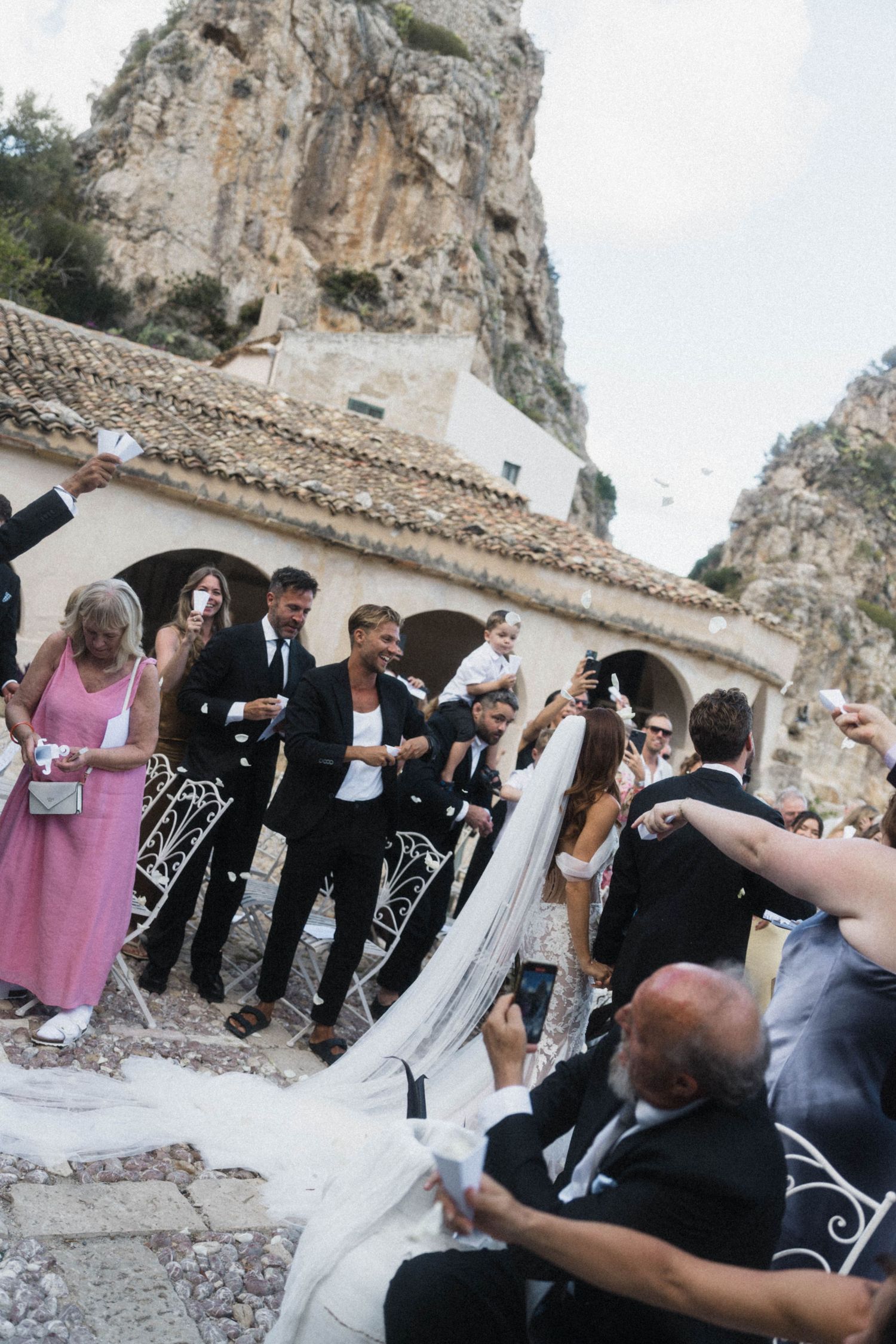 Wedding guests celebrate newlyweds with confetti near scenic rocky coastal venue.