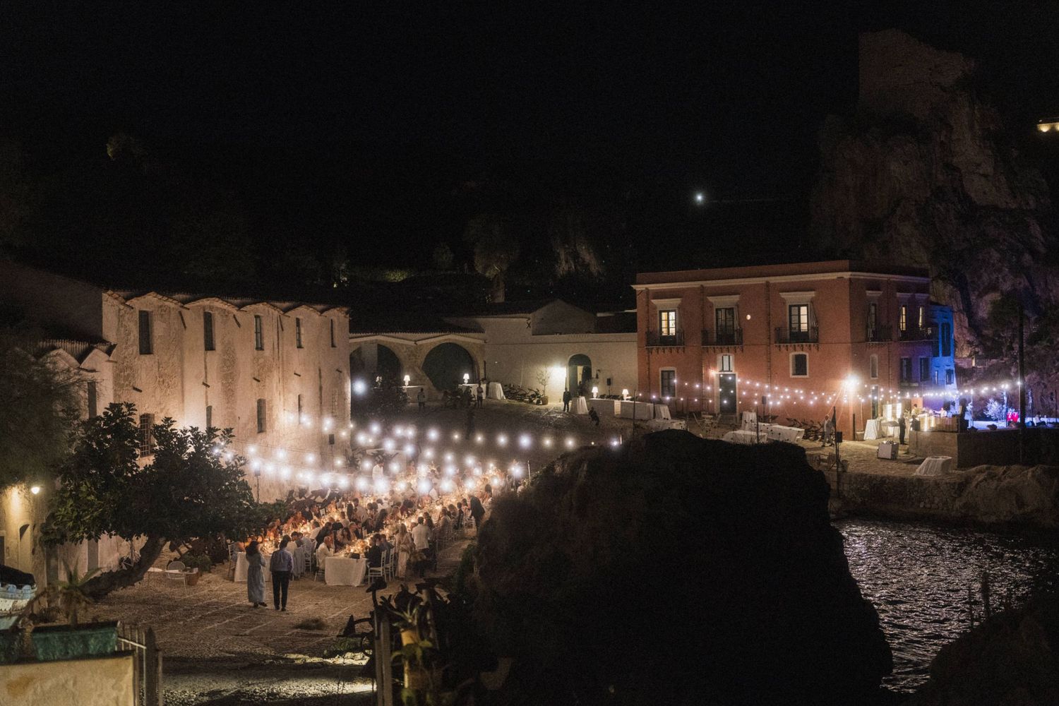 Breathtaking nighttime outdoor wedding reception with string lights set against historic stone buildings by the sea.