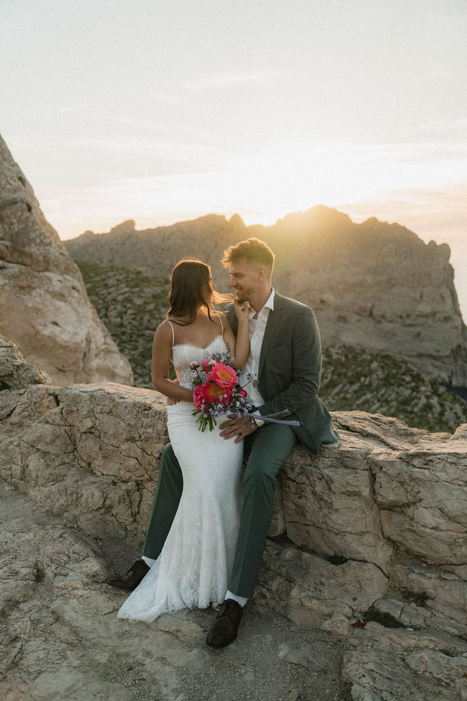 Bride and groom sit together on rocky ledge at golden sunset with mountain landscape backdrop, sharing an intimate moment.