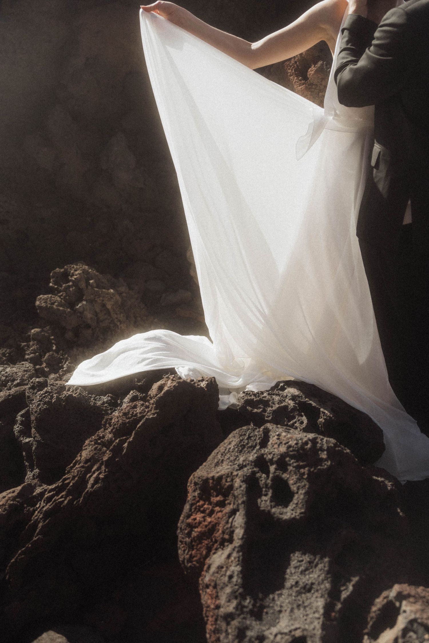 Bride in flowing white wedding dress standing on volcanic rocks, white fabric billowing dramatically in the wind.