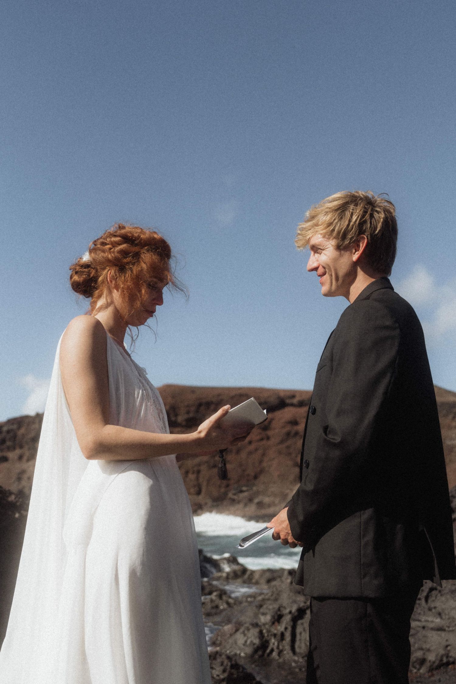 Bride reads vows to smiling groom in black suit on rocky coastal cliffside.