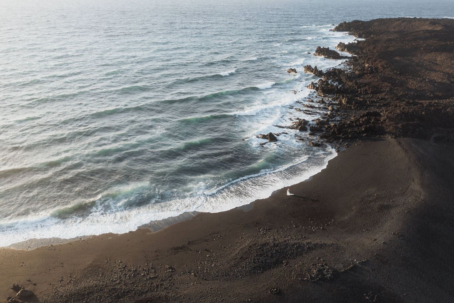 Aerial view of black sand beach with gentle ocean waves and rocky volcanic coastline.