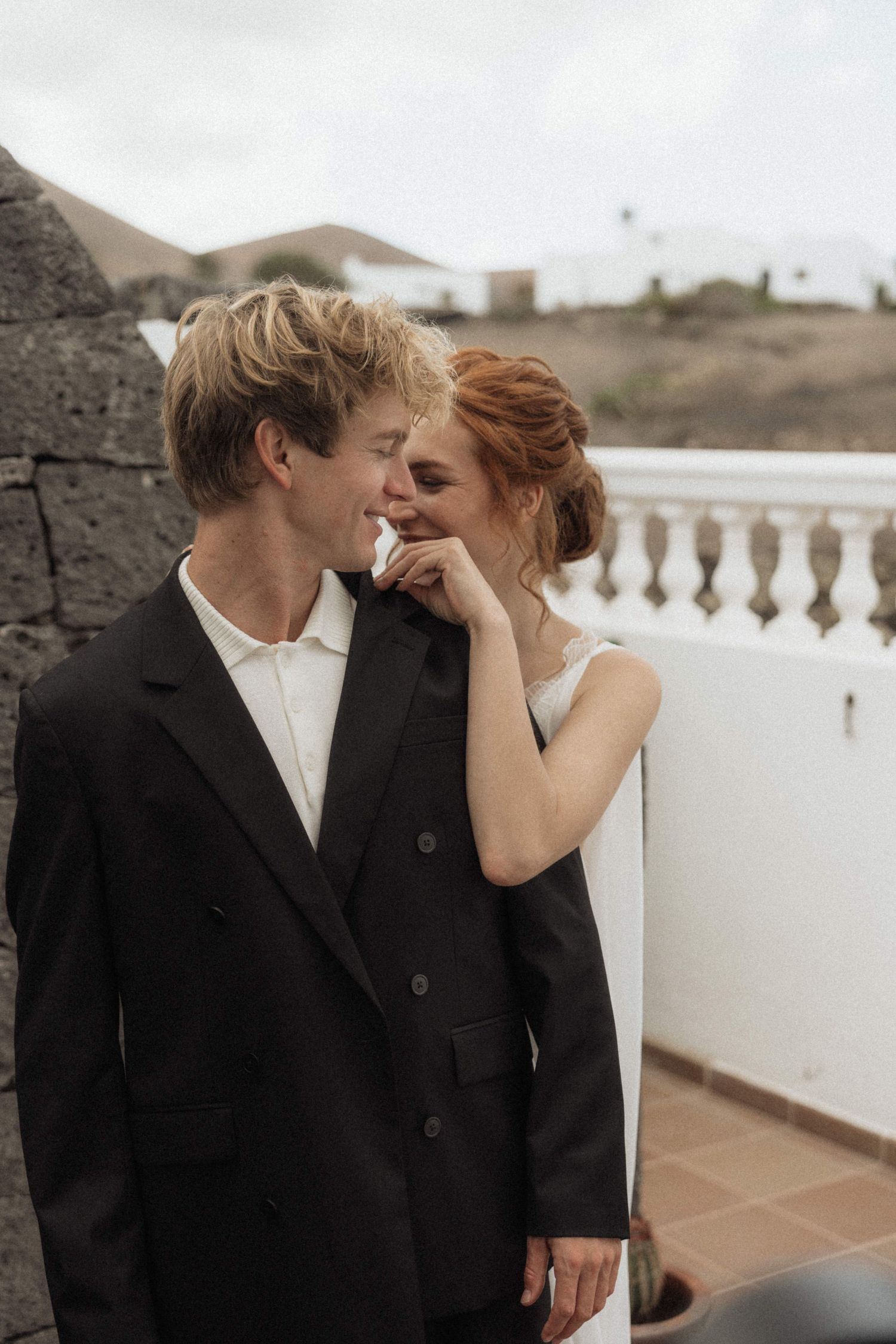 A smiling bride and groom share an intimate moment on a terrace with scenic mountain views behind them.