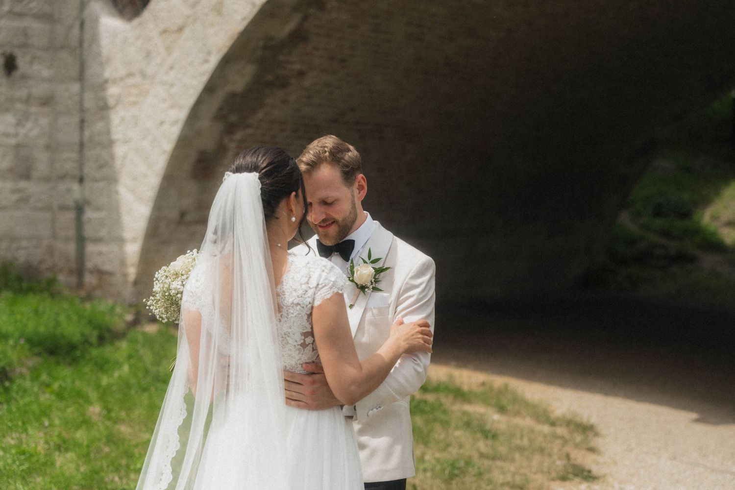 A bride and groom share a tender embrace outdoors near a stone arch, the bride wearing a veil and the groom in a gray suit.