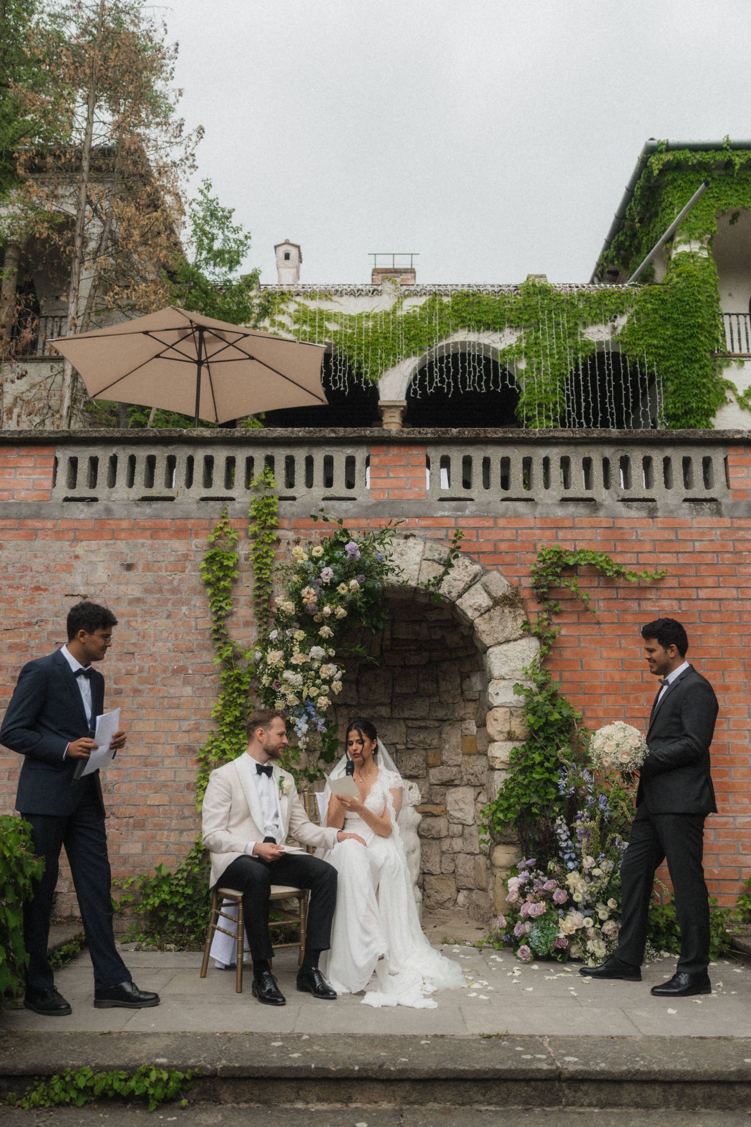 Bride and groom seated during outdoor wedding ceremony at brick wall venue with floral arch decor.