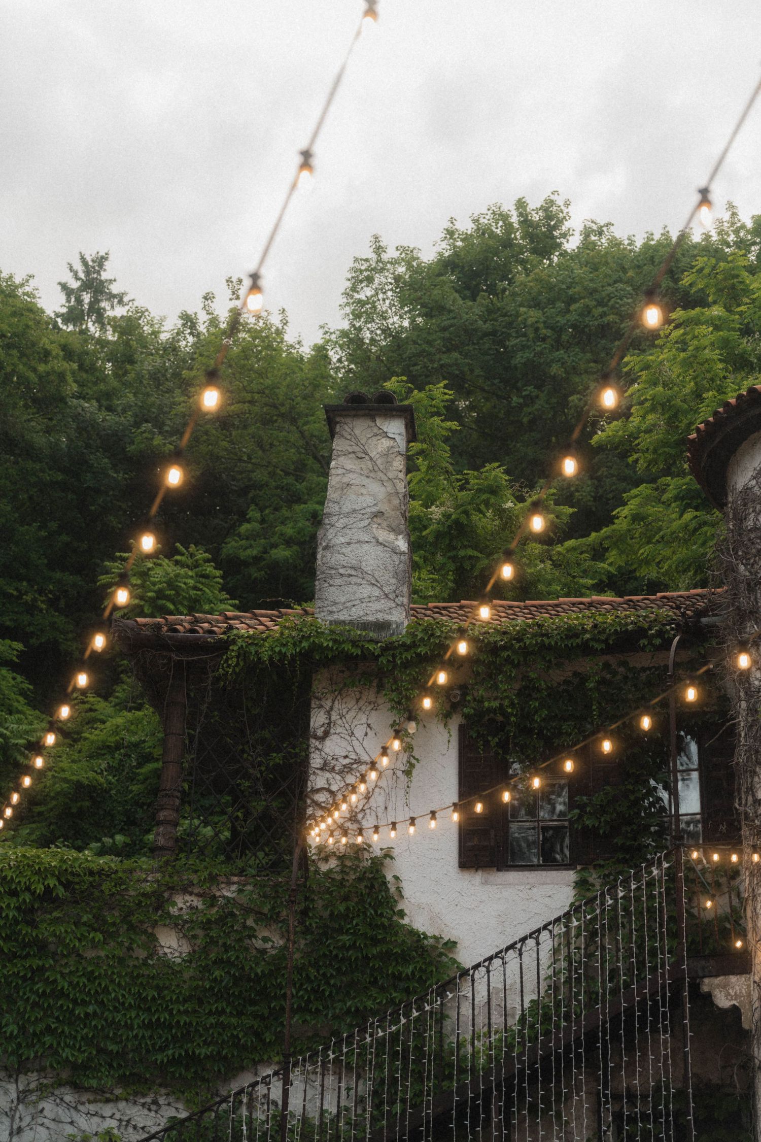 Rustic stone building covered in ivy with warm string lights glowing against an overcast evening sky.