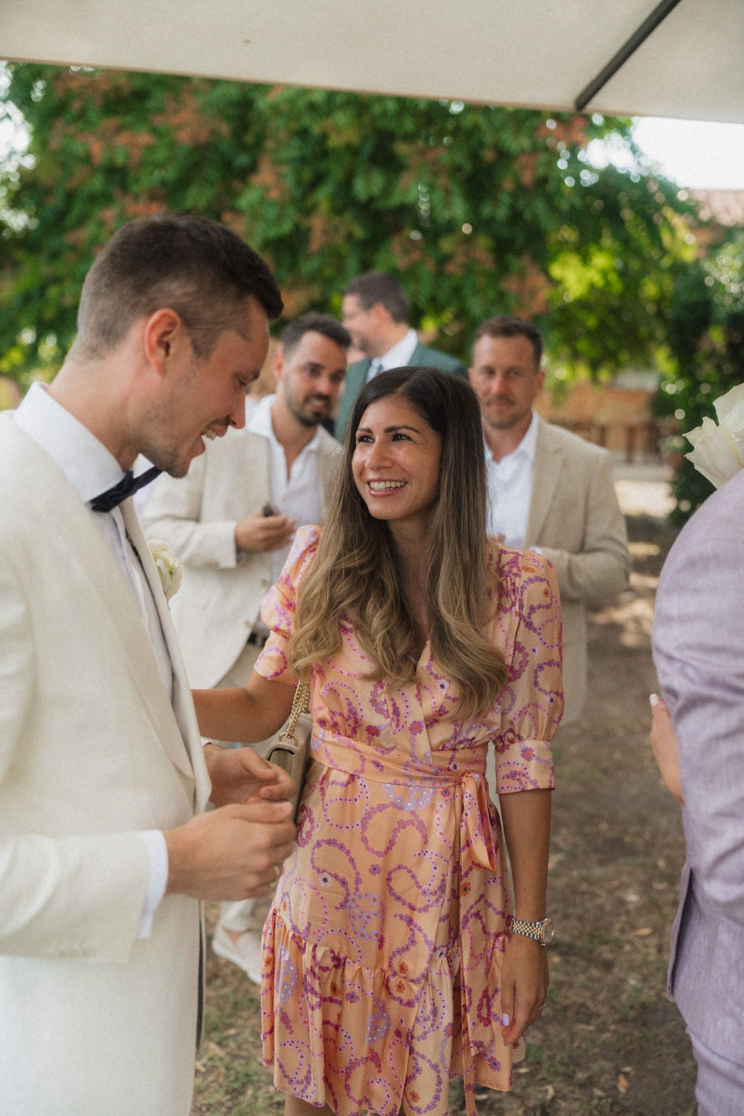 Groom in white tuxedo smiling with female wedding guest in pink floral dress at outdoor reception.