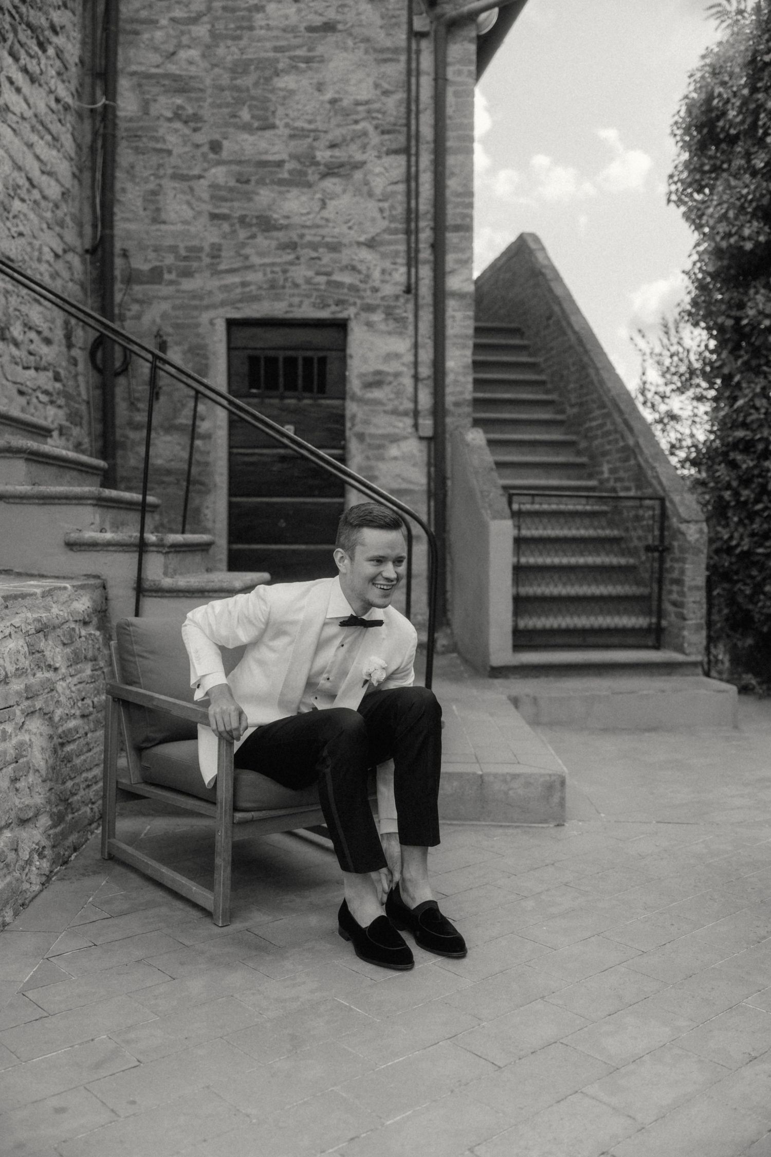 Groom in white shirt and bow tie sitting on chair outside rustic stone building, black and white wedding photo.