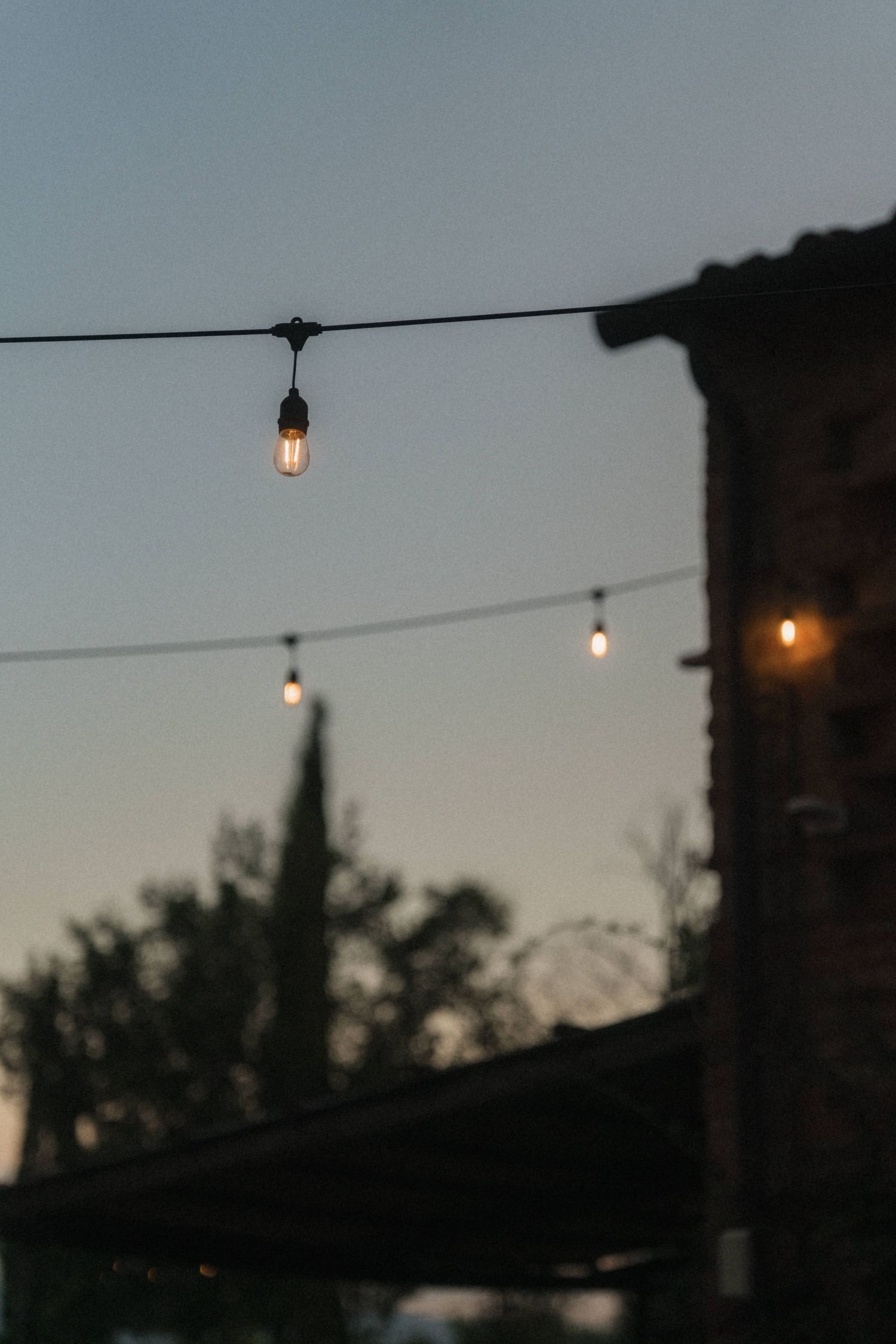 String of vintage Edison bulb lights hanging against a dusk sky near a rustic stone building outdoors.