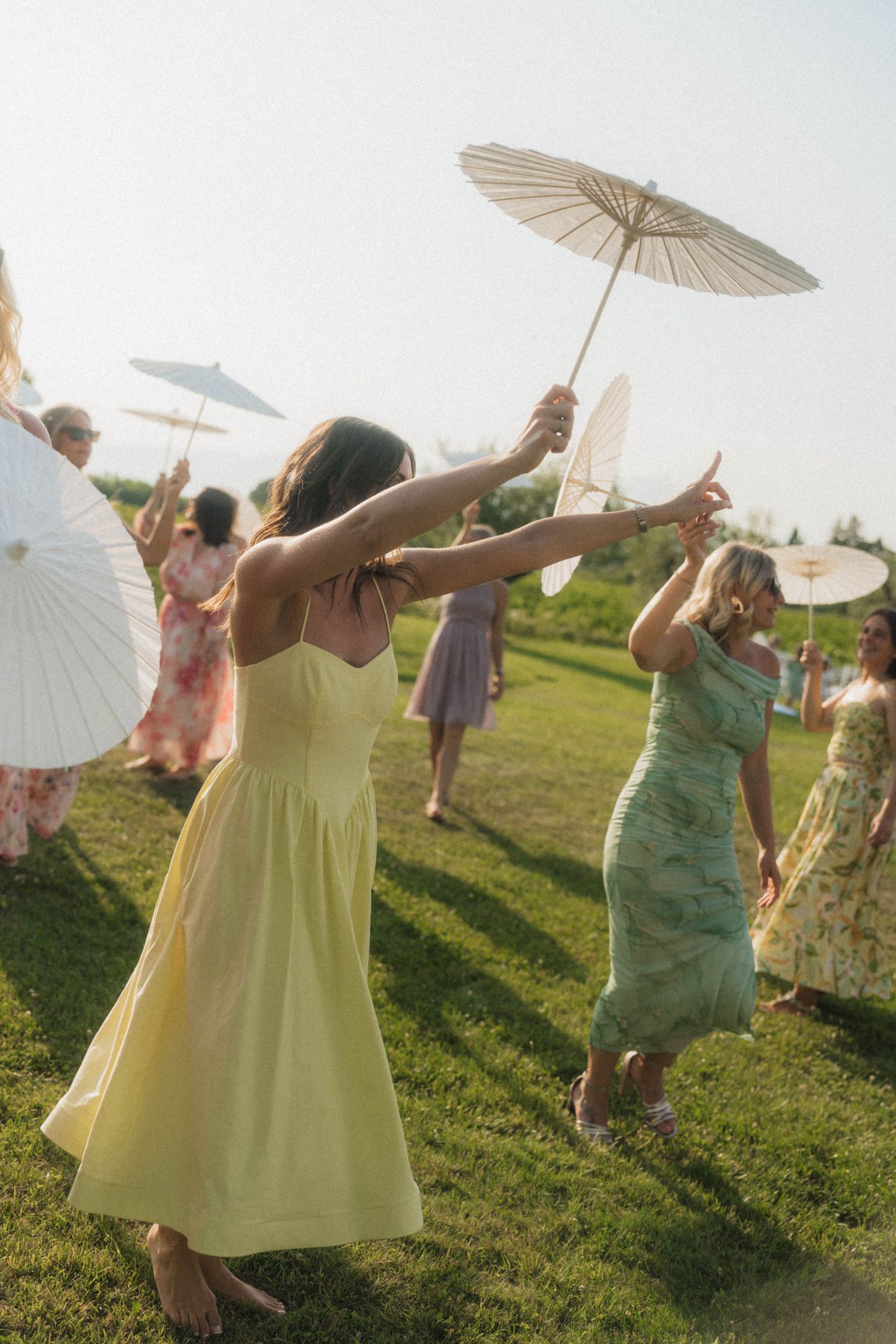 Wedding guests in colorful dresses dancing outdoors on grass while holding white paper parasols.