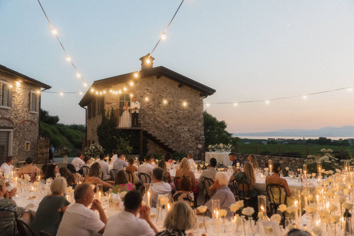 Romantic outdoor wedding reception at dusk with string lights and stone building backdrop.