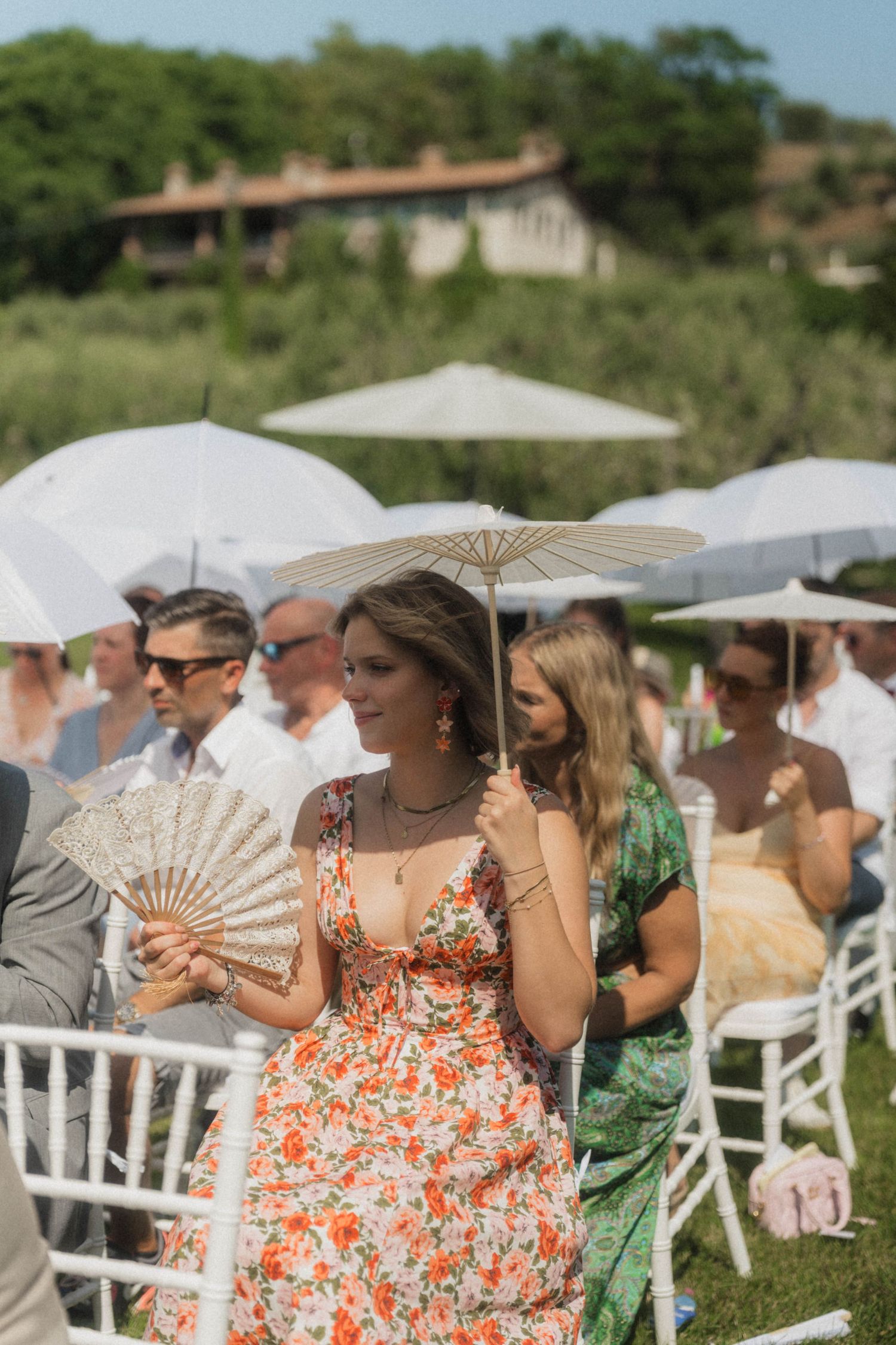 Wedding guest in orange floral dress holding fan and parasol at outdoor summer ceremony.