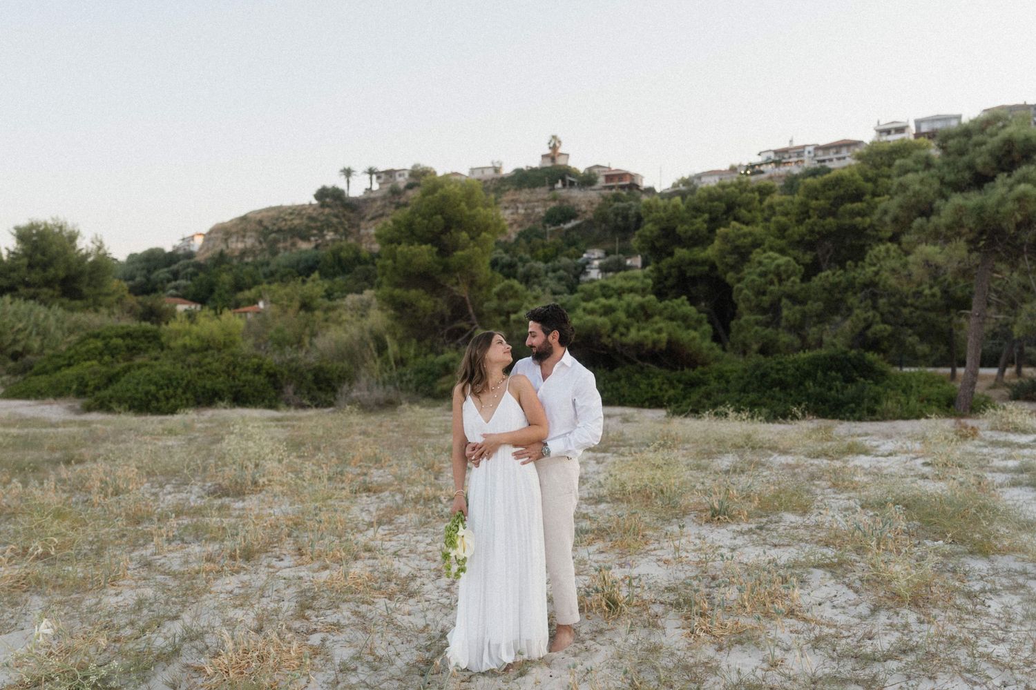 Couple in white wedding attire sharing a romantic moment in a scenic outdoor field with lush greenery and hillside behind them.