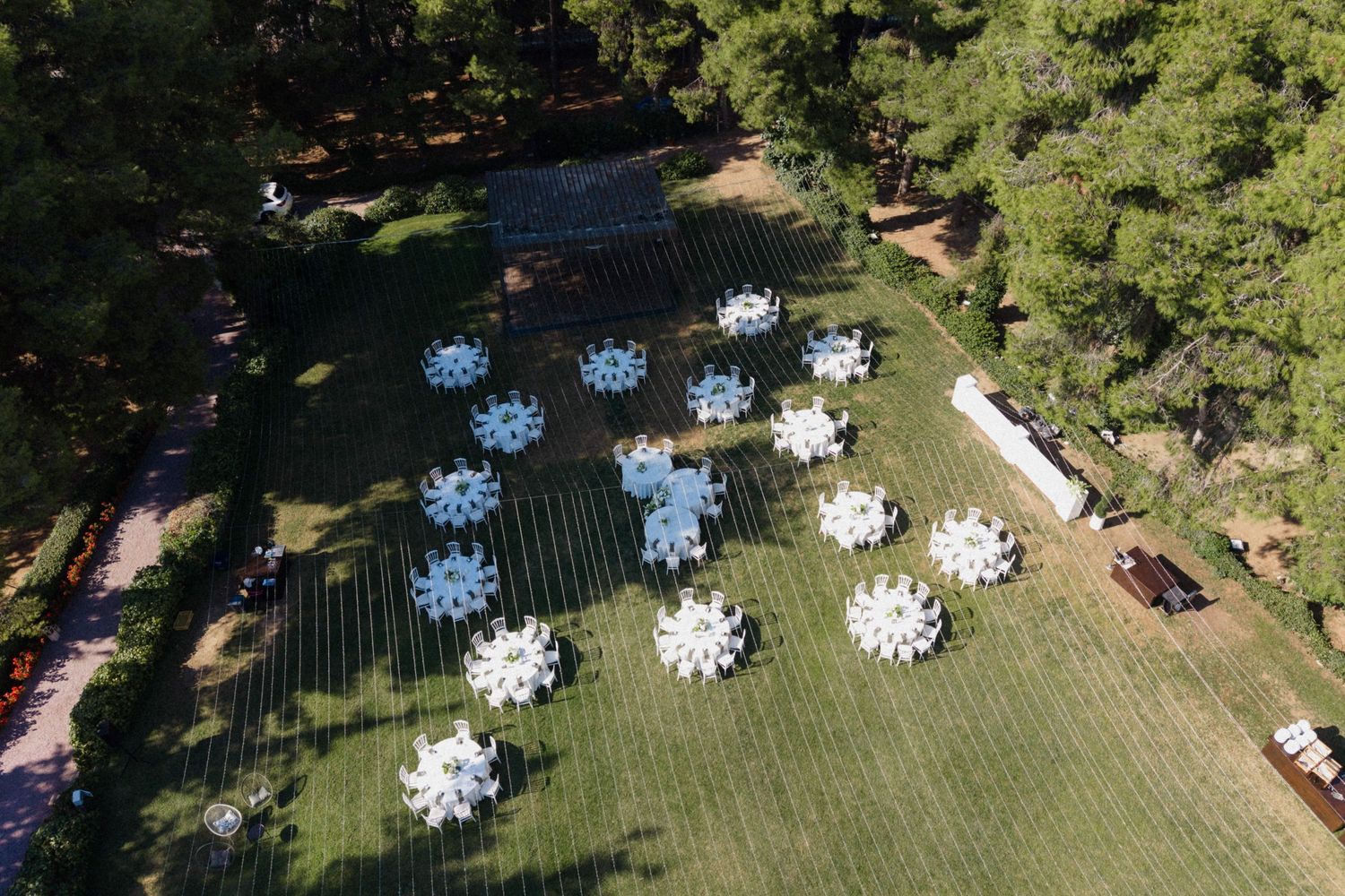 Aerial view of outdoor event setup with round white-clothed tables arranged on green lawn surrounded by tall pine trees.