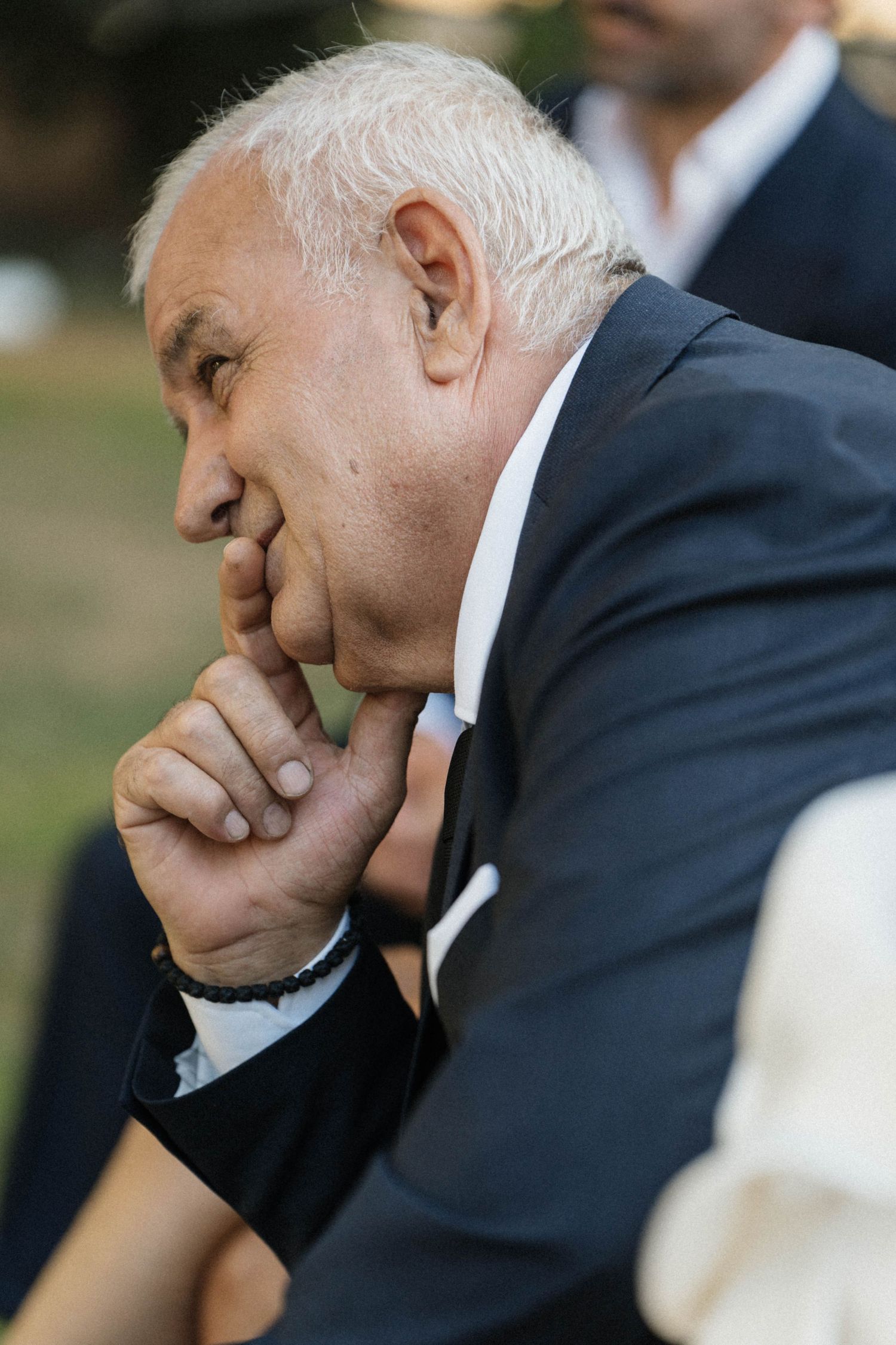 Elderly man in dark suit sits thoughtfully with hand raised to his chin during an outdoor wedding ceremony.