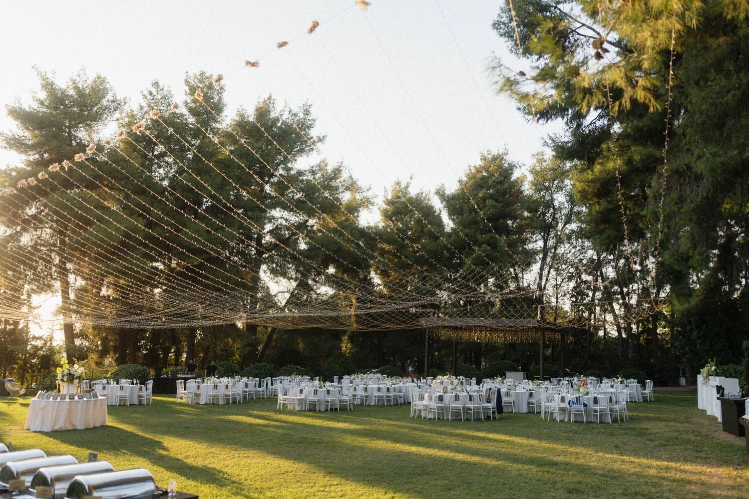 Outdoor wedding ceremony setup with white chairs on green lawn, string lights overhead, surrounded by tall trees at golden hour.