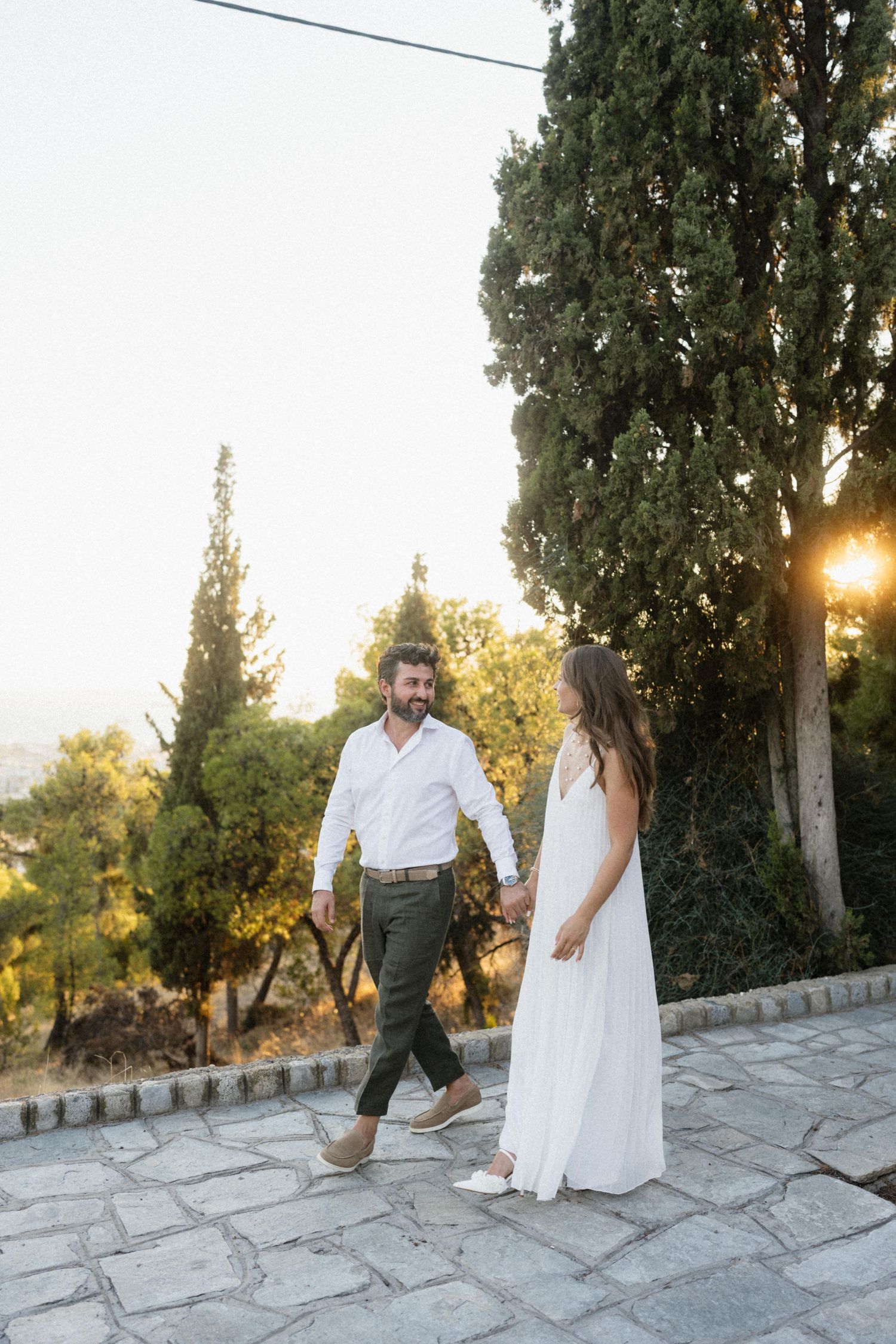 Couple in white attire holding hands and walking on stone path at sunset with cypress trees in background.