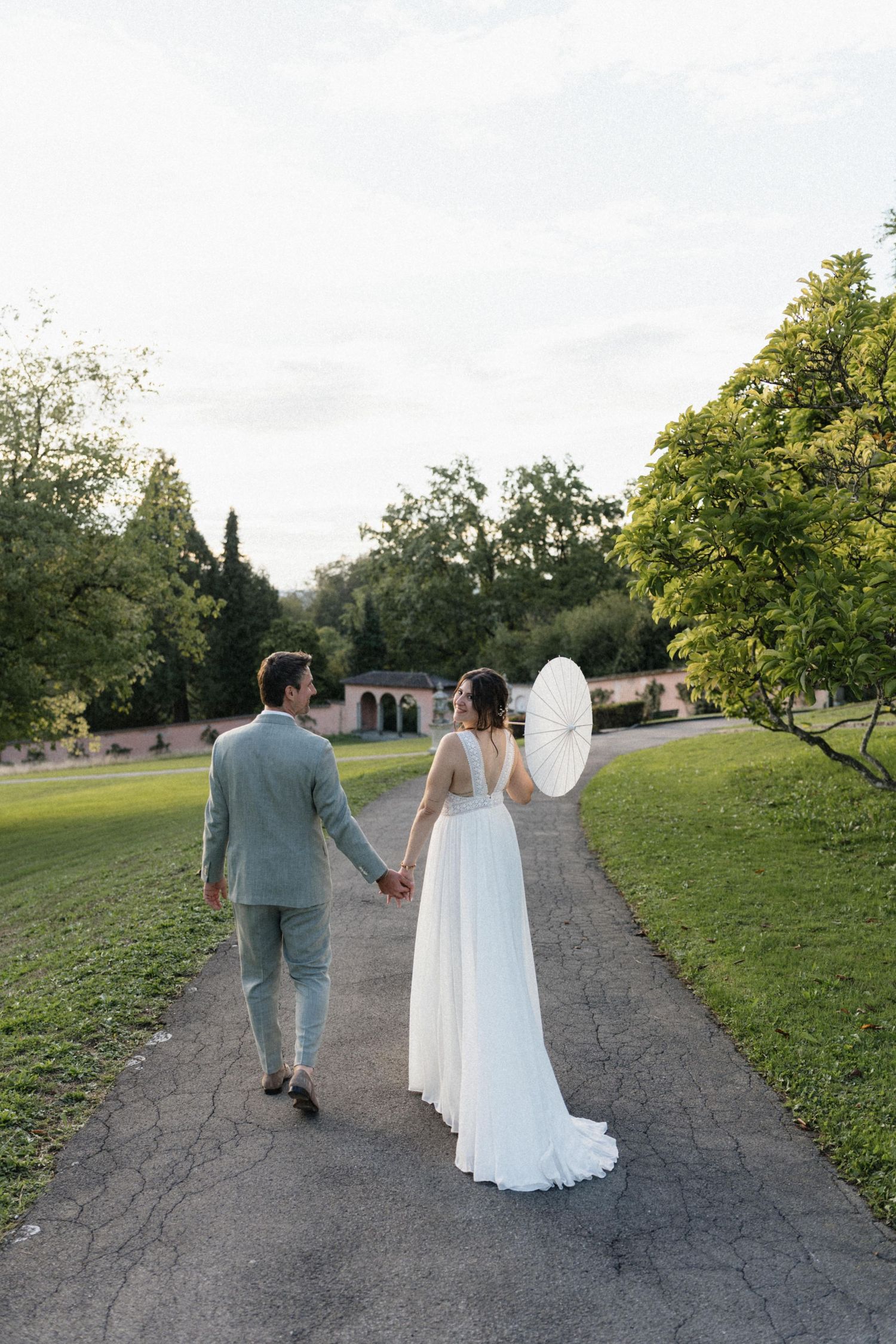 Bride in white gown holding parasol and groom in gray suit walking hand in hand in park.