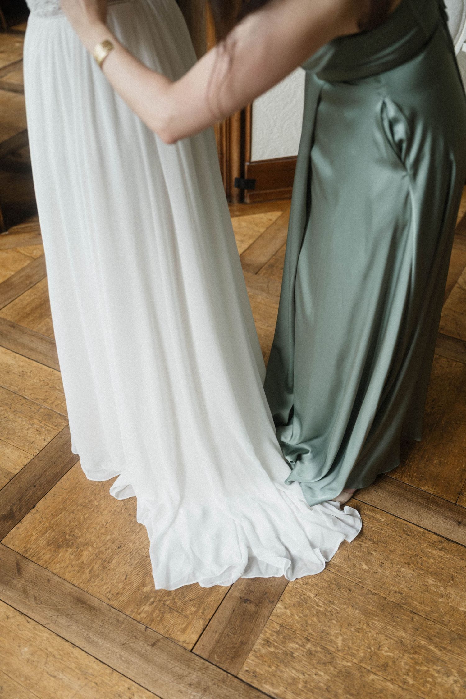 Bride in white chiffon gown being helped by bridesmaid in sage green satin dress on hardwood floor.