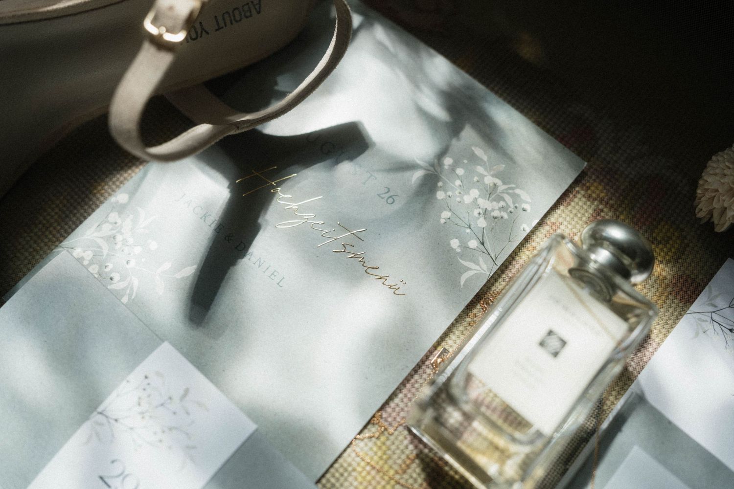 Jo Malone perfume bottle beside a floral wedding invitation on a light blue silk ribbon flat lay.
