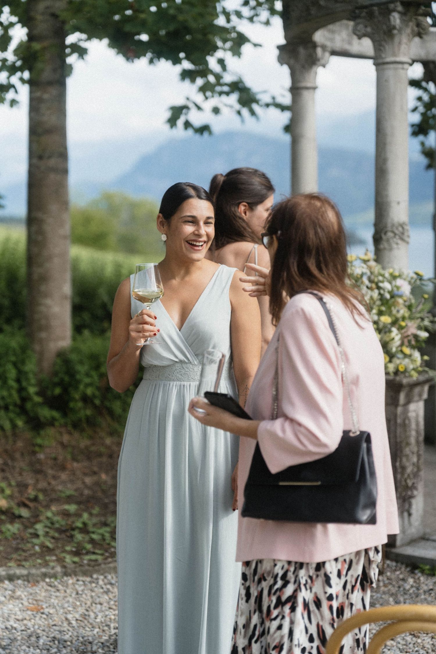 Bridesmaid in light blue gown laughing and holding white wine glass while chatting with guests at an outdoor wedding reception.
