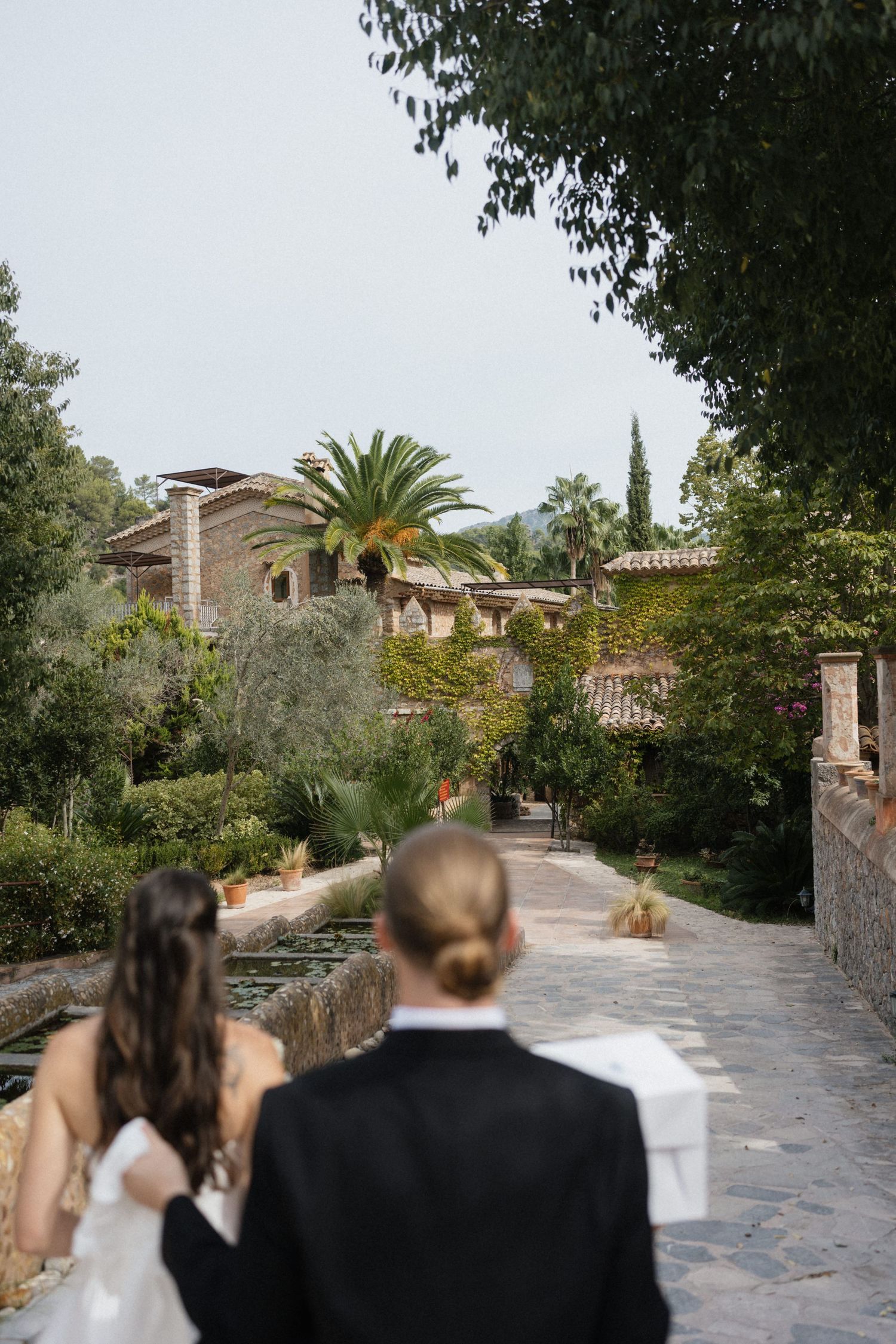 Bride and groom walk away along a stone path toward a rustic Mediterranean villa surrounded by palm trees and lush greenery.