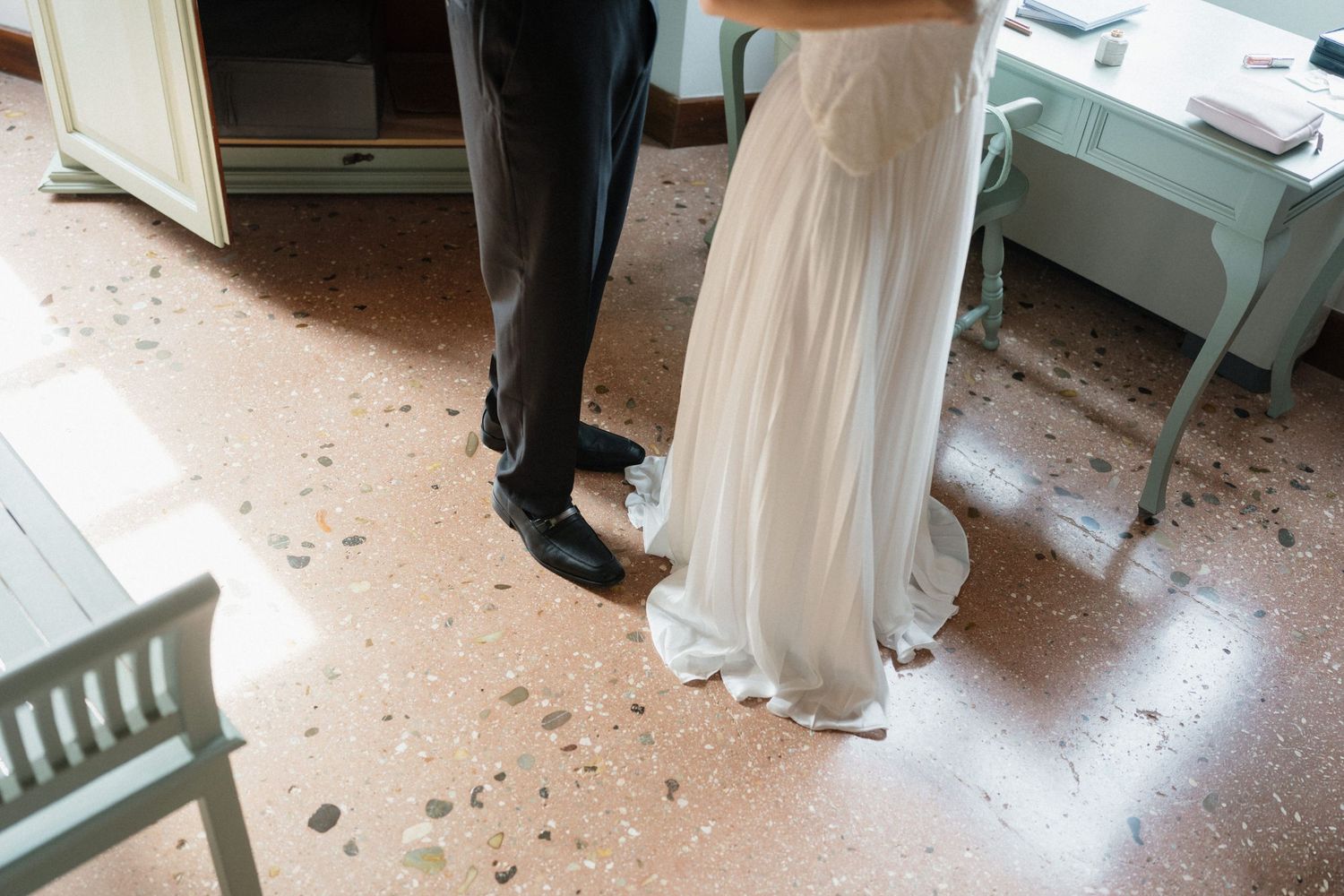 Close-up of a groom in dark trousers and black shoes standing beside a bride in a flowing white wedding dress on a speckled terrazzo floor.