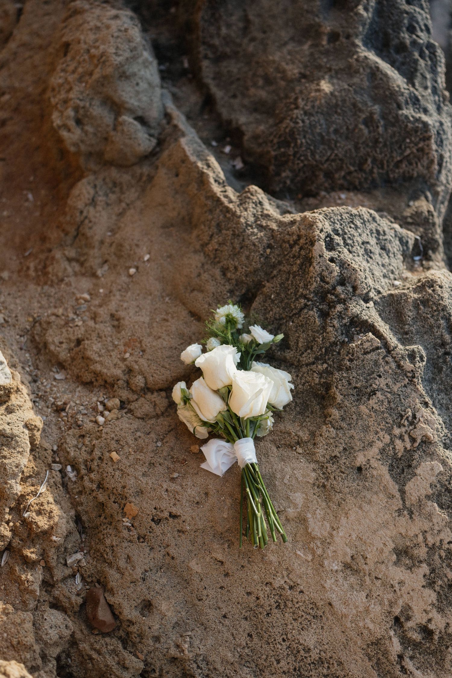 A small bridal bouquet of white roses and greenery tied with ribbon rests on a textured, weathered rock surface.