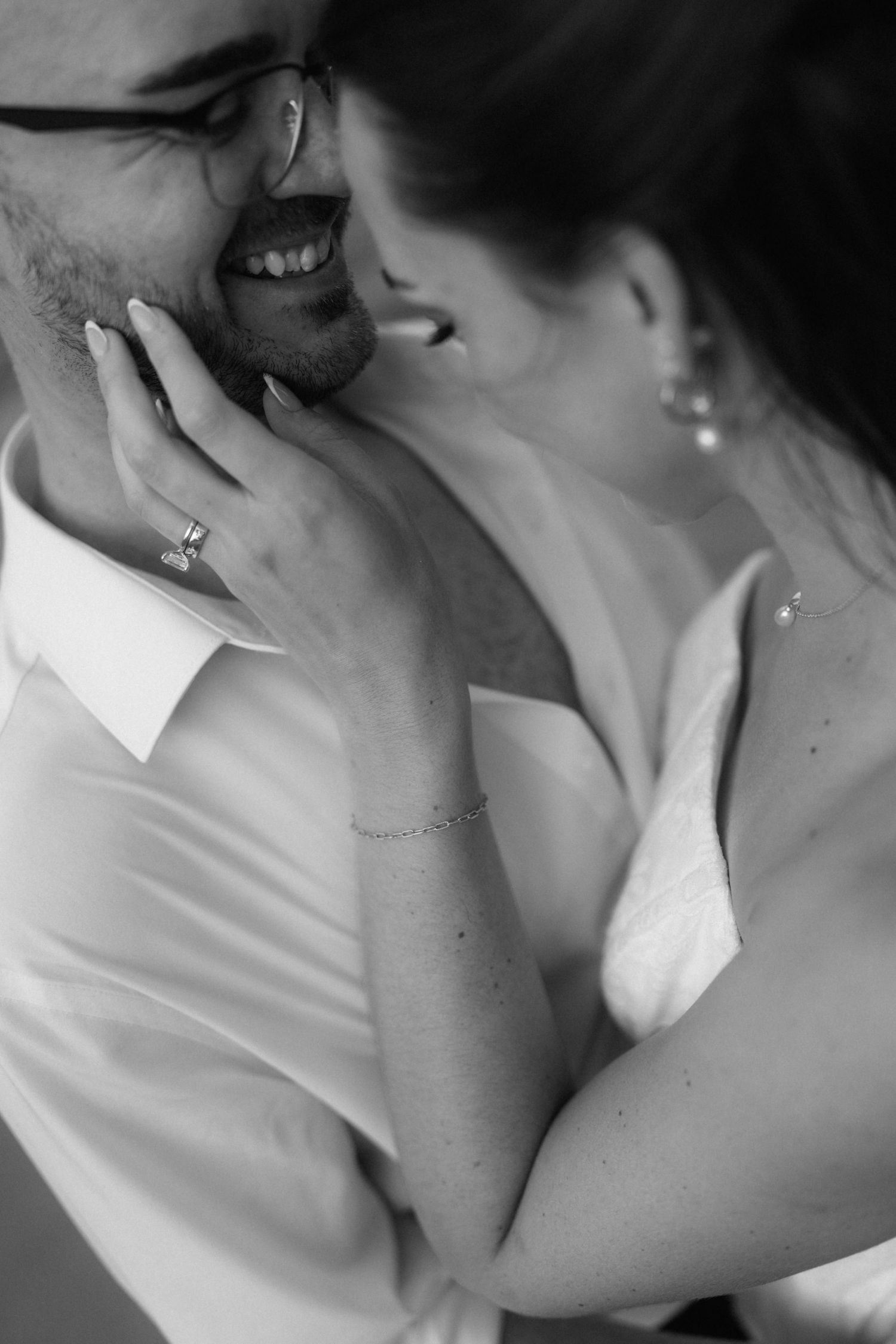 Black and white close-up of a smiling couple embracing, with her ringed hand gently touching his bearded cheek in an intimate moment.