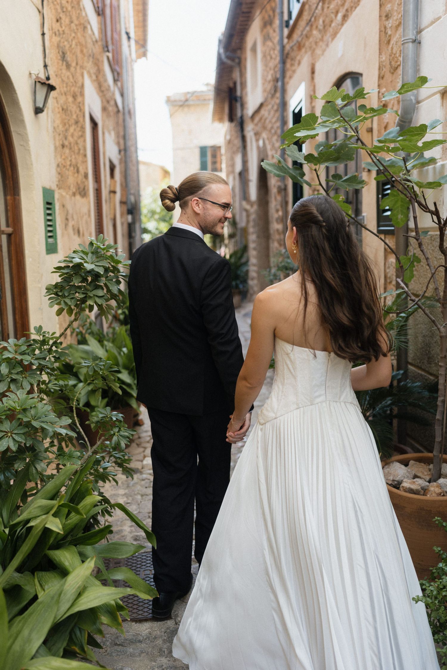 Bride in a white strapless gown and groom in a black suit walk hand in hand down a narrow stone alley lined with plants.