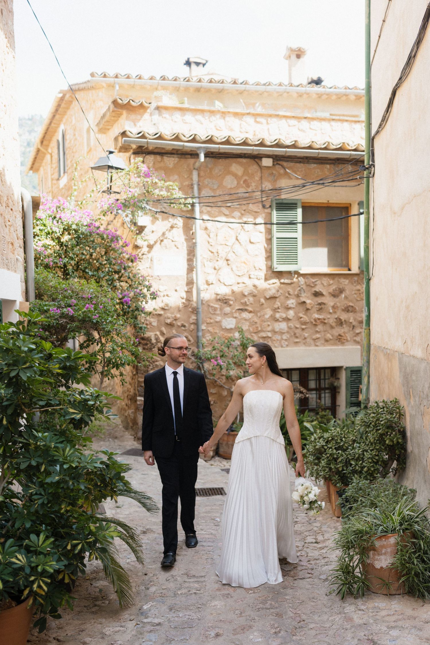 Bride in a white strapless gown and groom in a black suit hold hands while walking down a charming stone alley lined with plants.