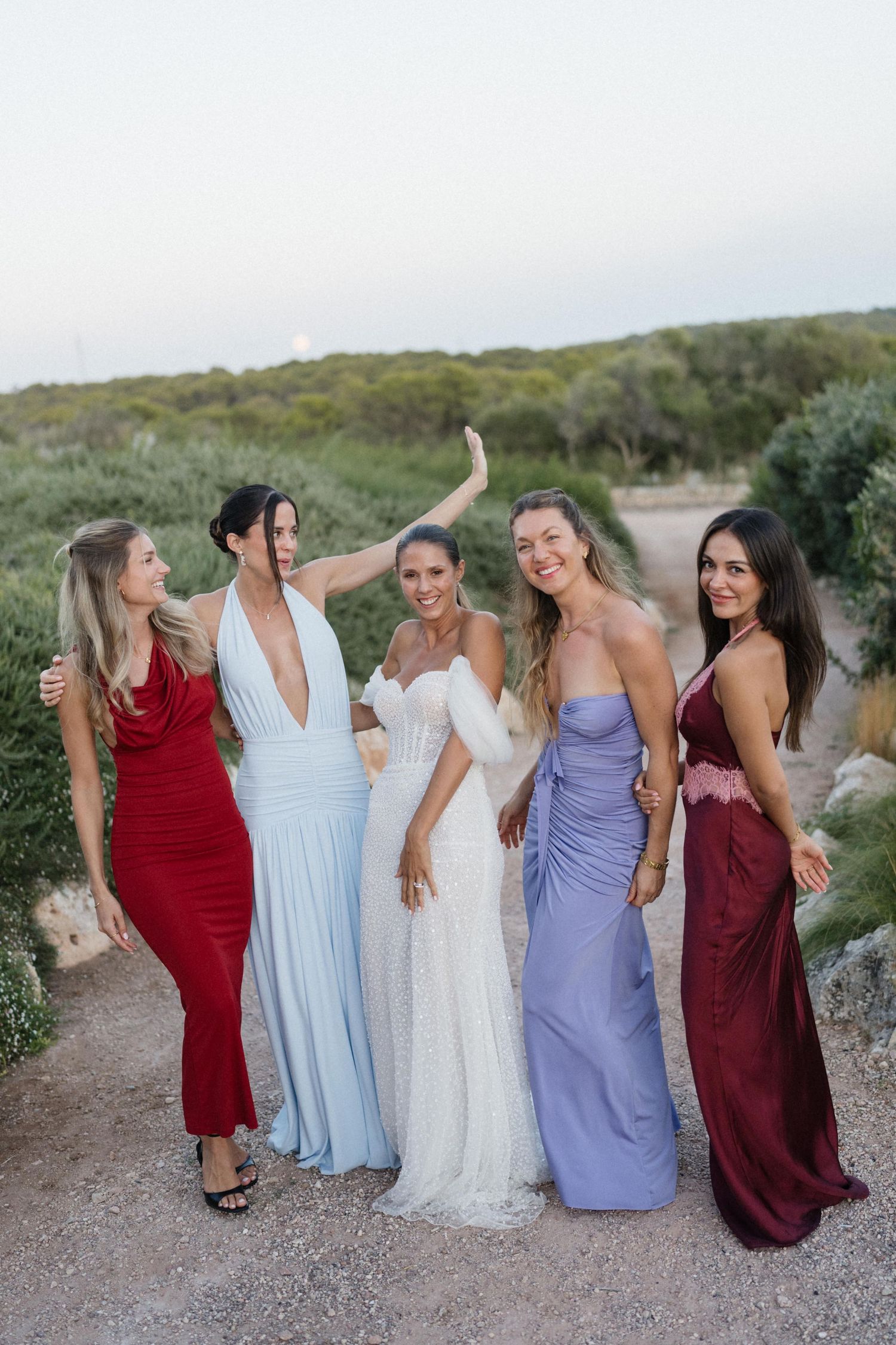 Five women in elegant red, blue, white, lavender and burgundy gowns pose together on a scenic dirt path surrounded by greenery at dusk.