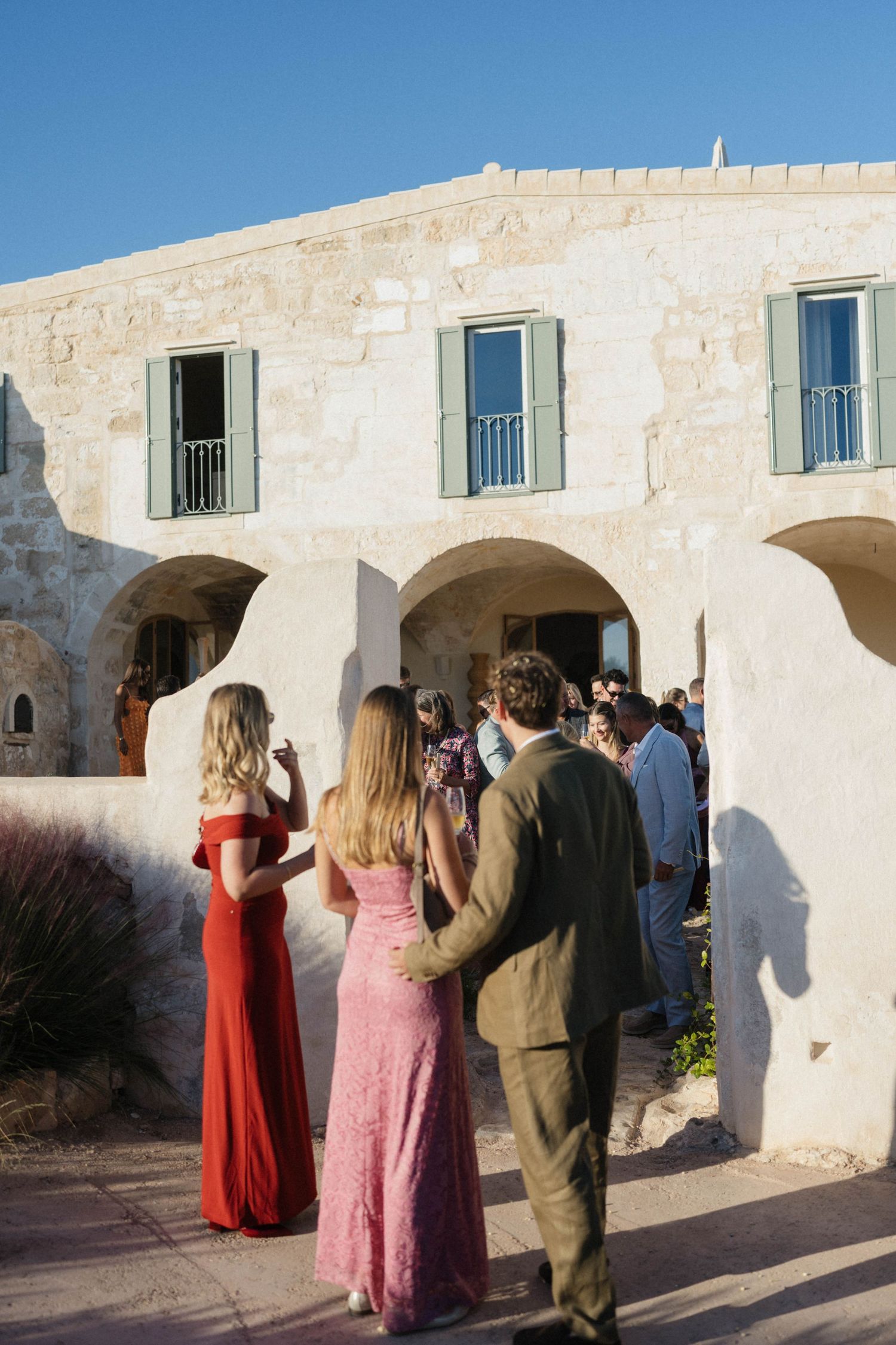 Wedding guests in red, pink and olive outfits mingle outside a rustic stone villa with green shutters at golden hour.