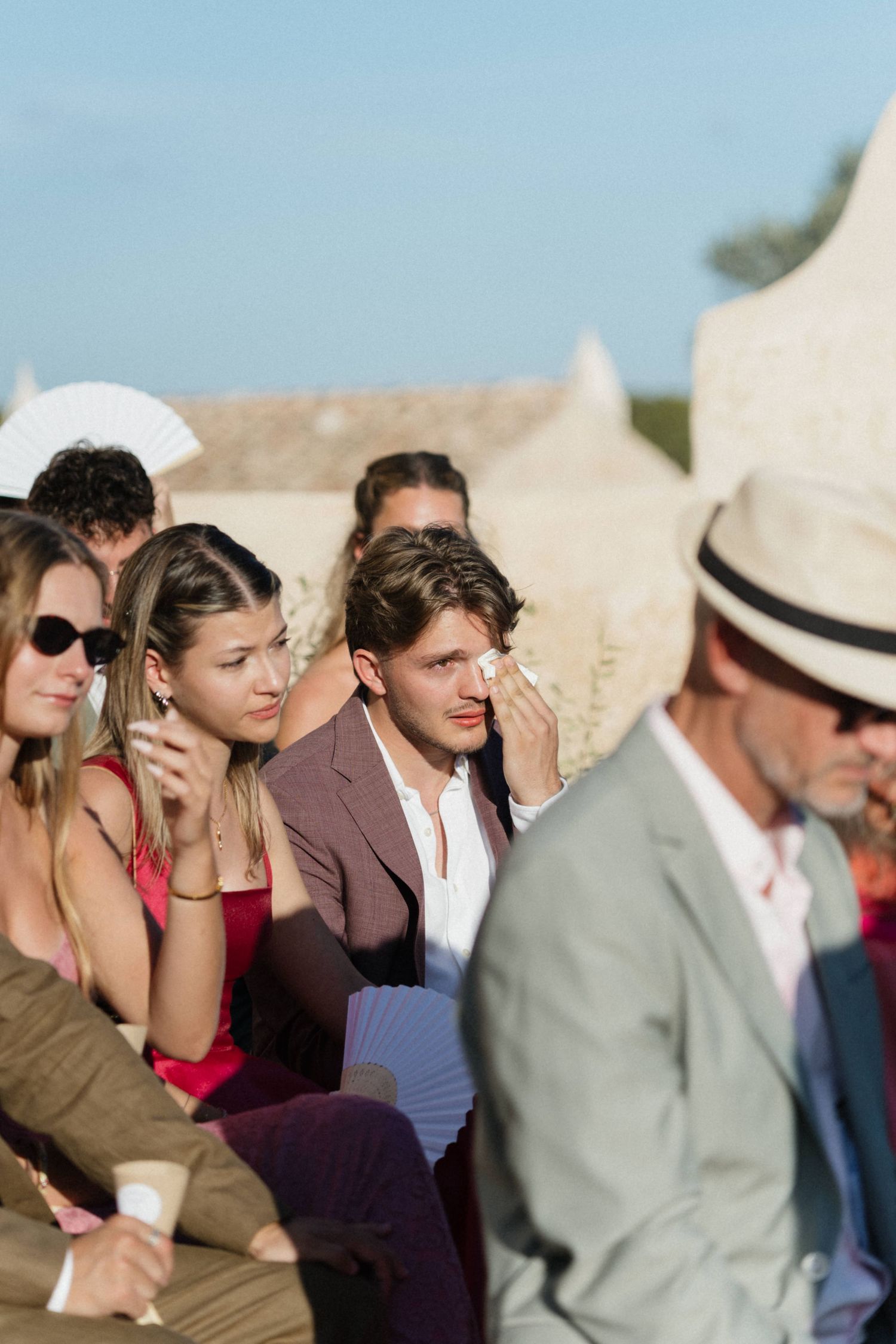 A guest in a burgundy blazer wipes away tears during an outdoor wedding ceremony as nearby guests watch.