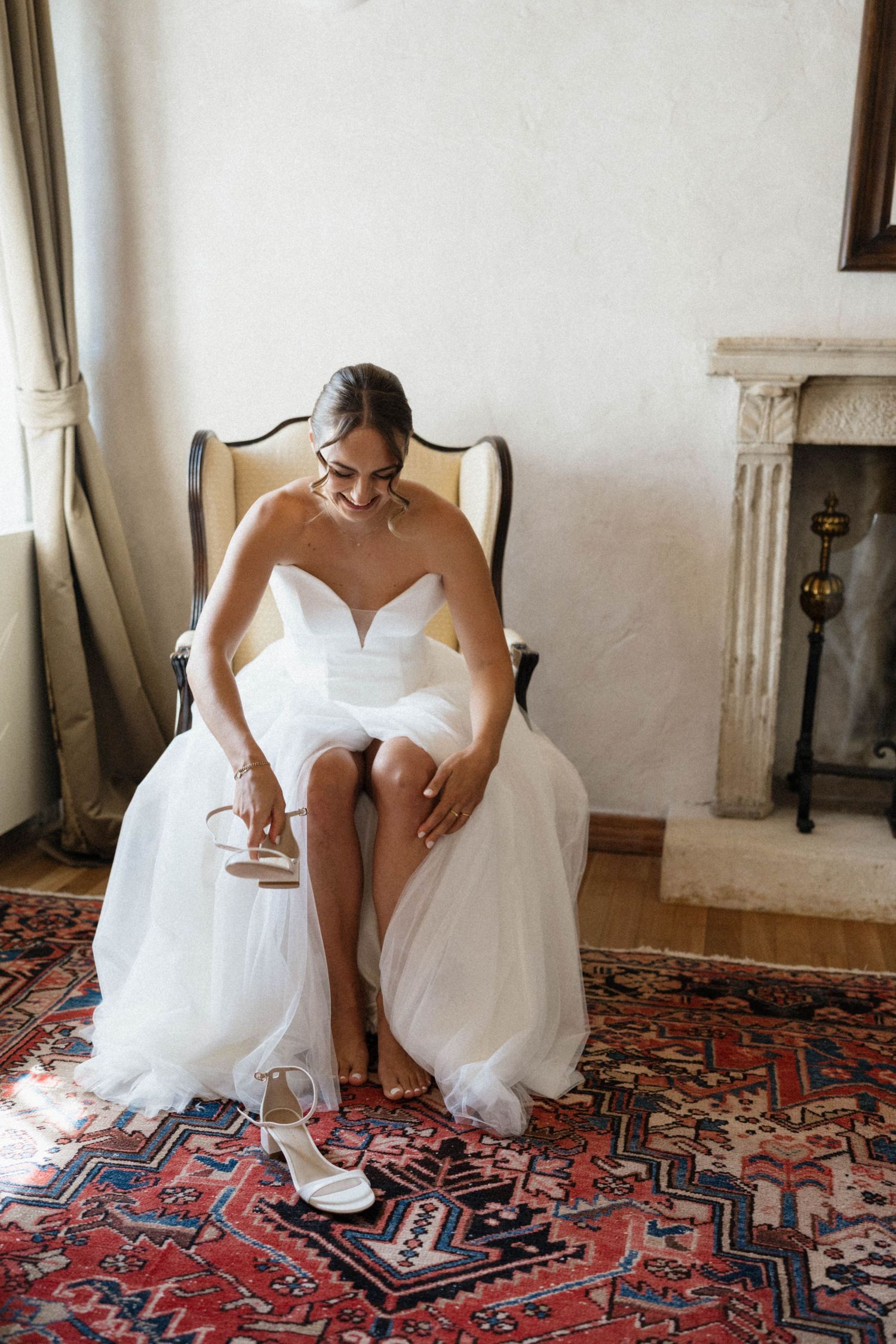 Bride in strapless white tulle gown putting on heels while seated in elegant chair on ornate rug.