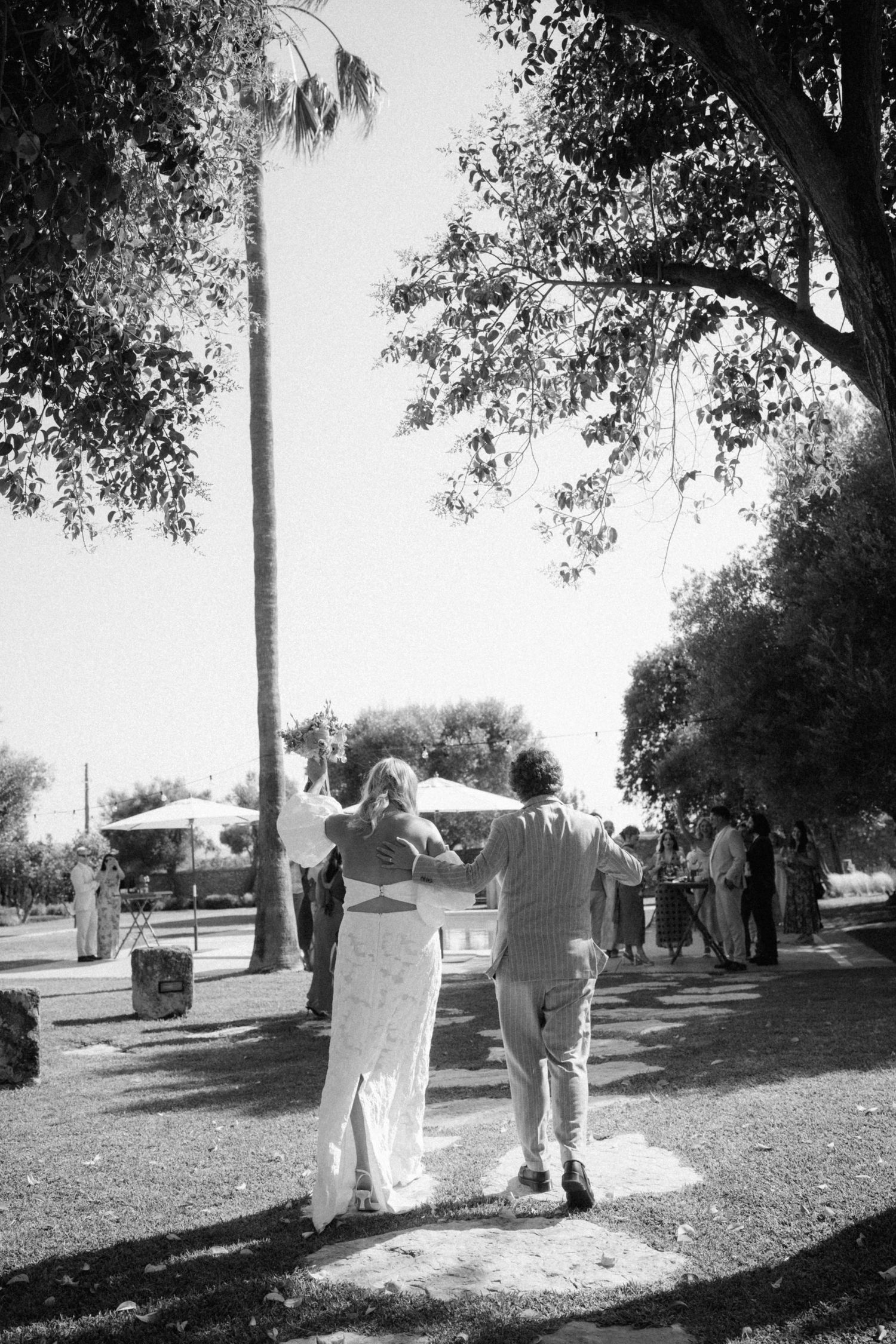 Black and white photo of bride and groom walking hand in hand through a tree-lined outdoor wedding venue.