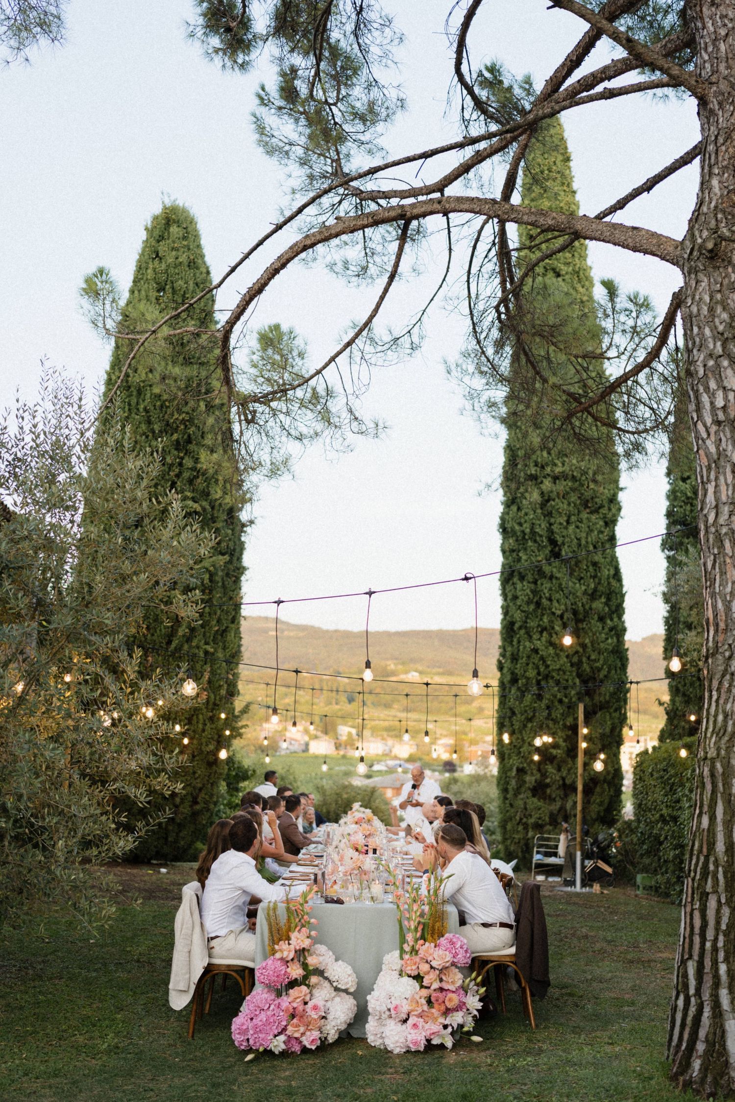 Outdoor wedding reception dinner under string lights with pink floral centerpieces, cypress trees, and rolling hills at dusk.