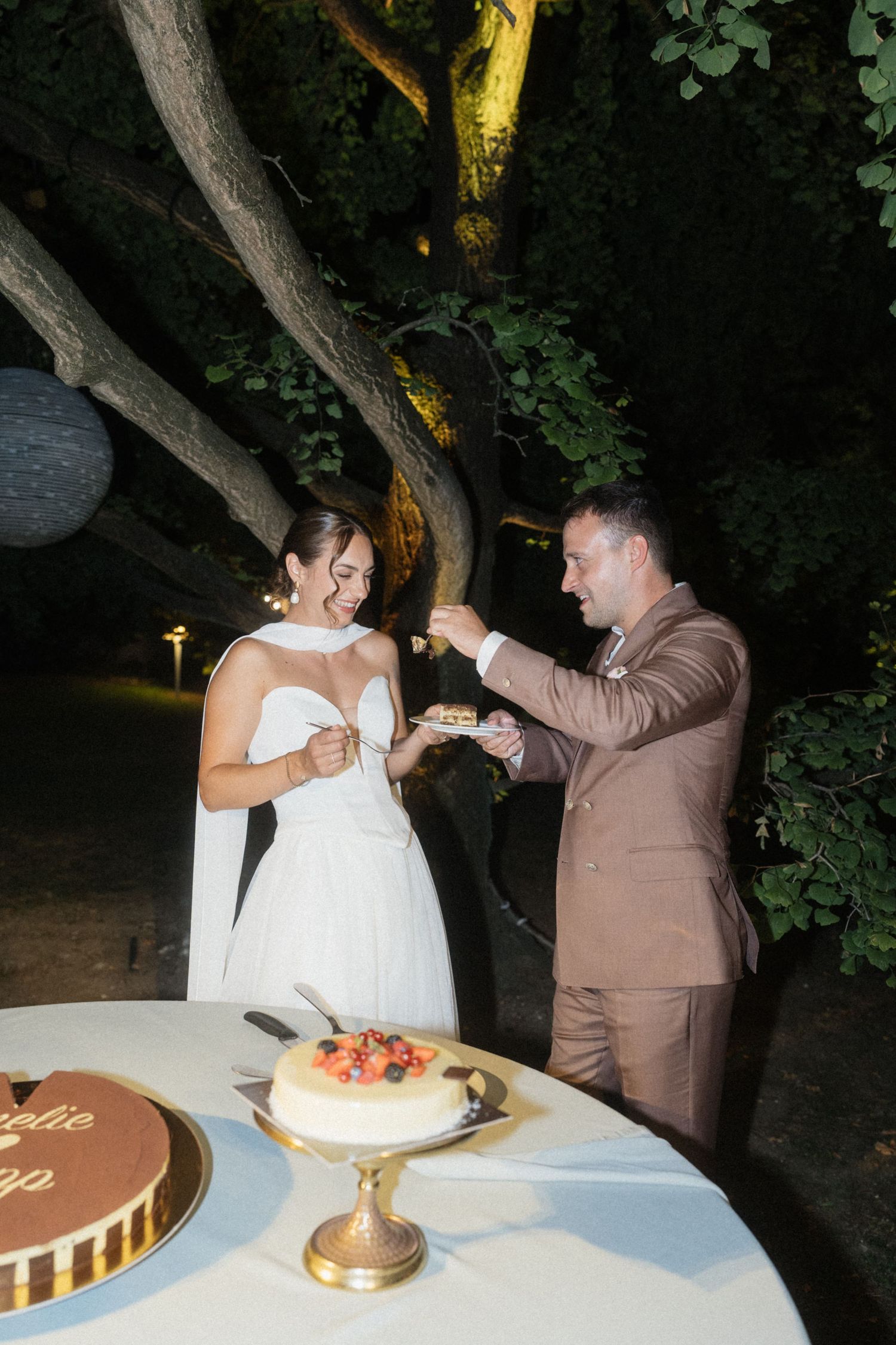 Bride and groom sharing wedding cake outdoors at night under trees, wearing white dress and brown suit.