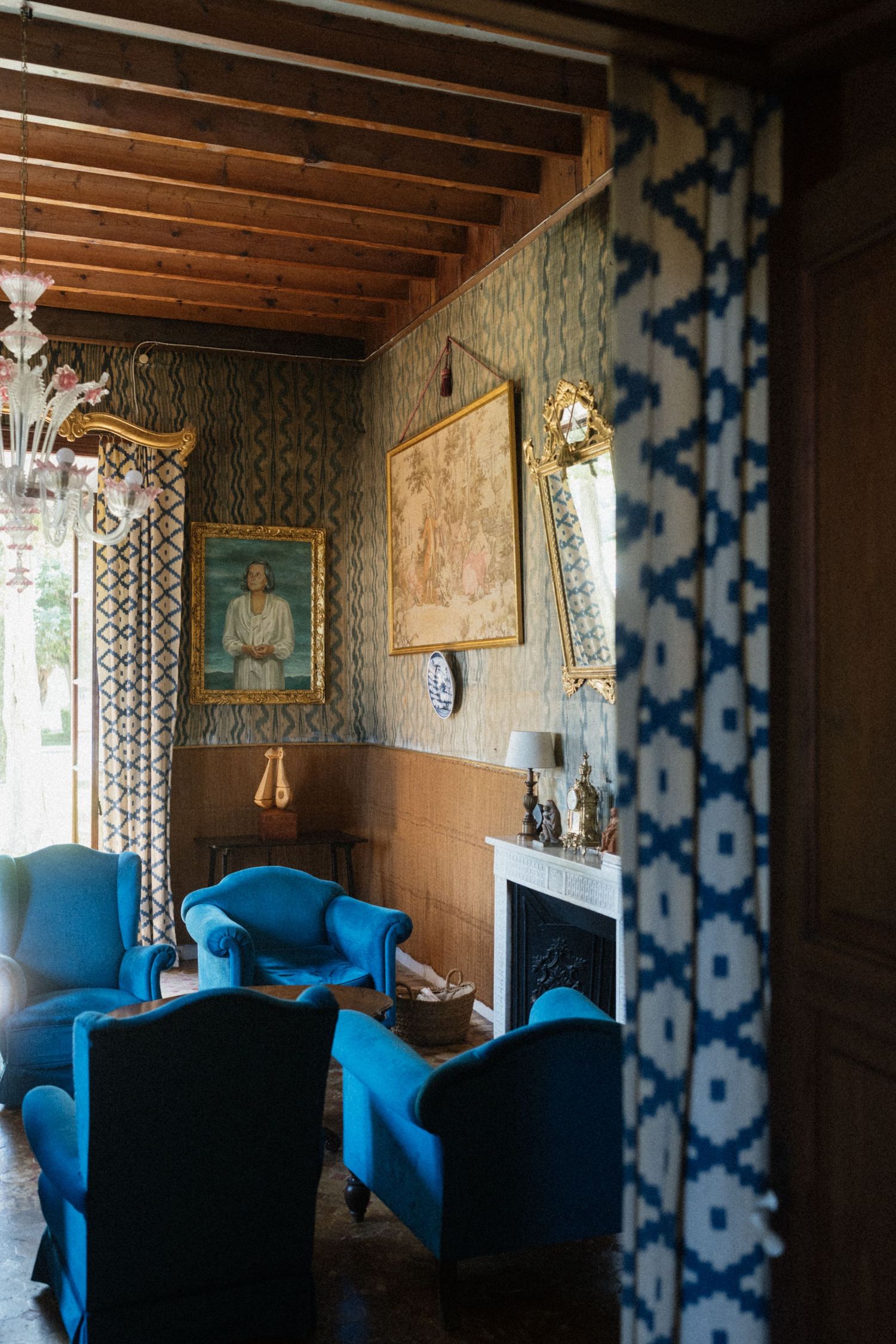 Elegant living room with vibrant blue armchairs, ornate gold mirror, portrait painting, patterned wallpaper, and wooden beam ceiling.