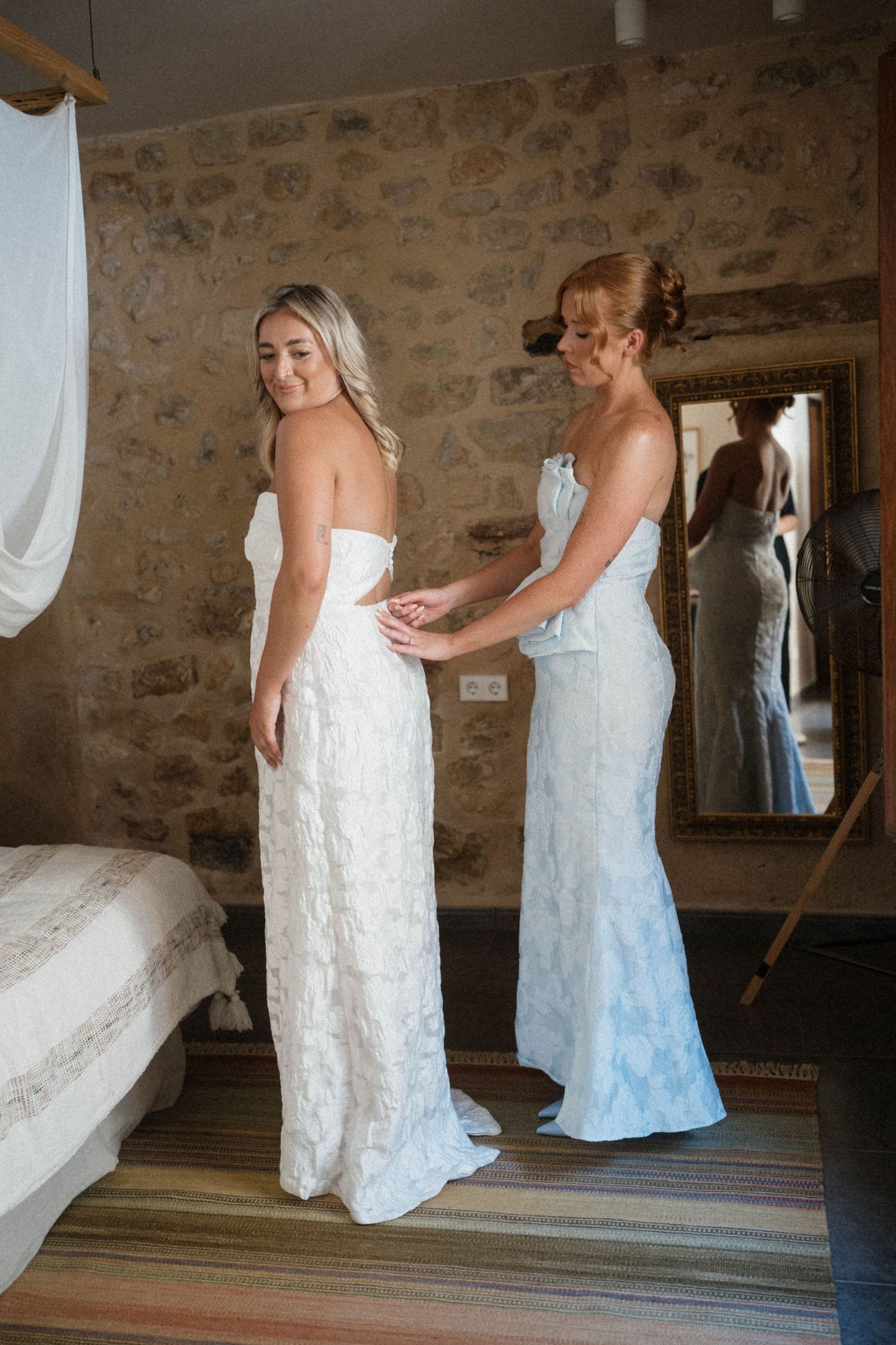Bridesmaid helping bride fasten her white lace wedding dress in a rustic stone-walled bedroom.