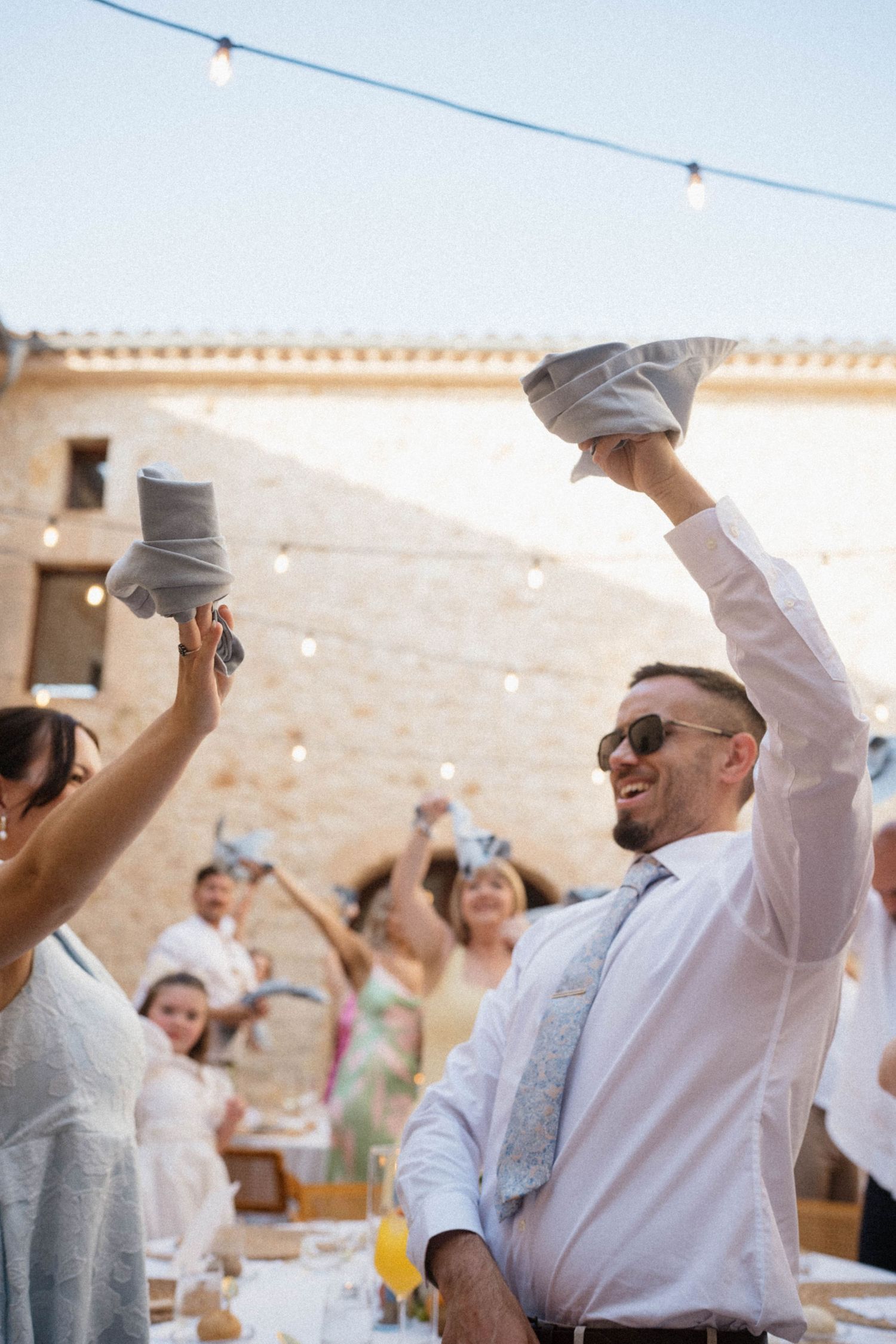 Smiling man in sunglasses raises napkin at outdoor wedding reception with string lights and guests celebrating.